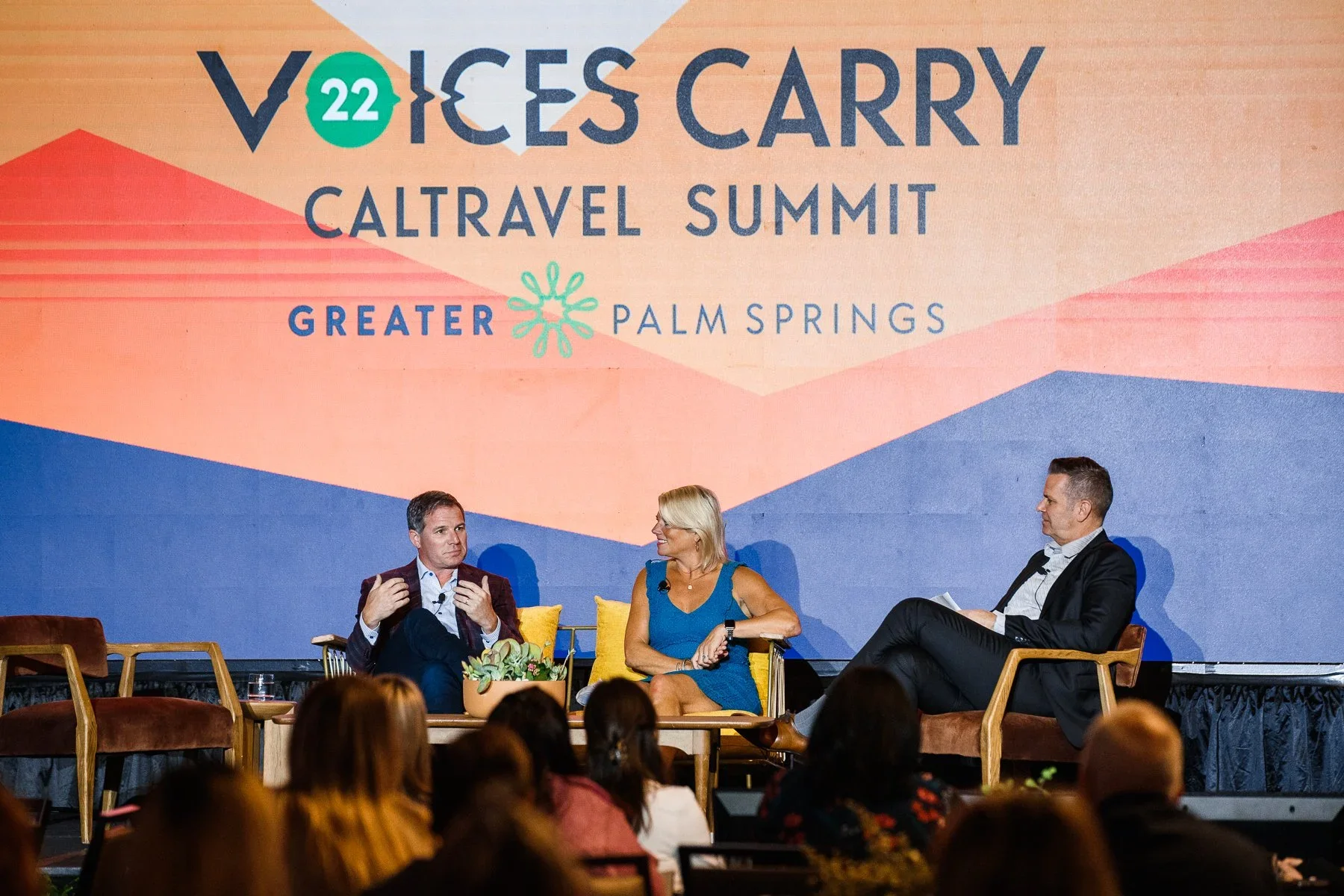 A panel discussion at the Voices Carry CalTravel Summit in Greater Palm Springs, with three people seated on stage, two men and one woman, engaging in conversation, with an audience watching.