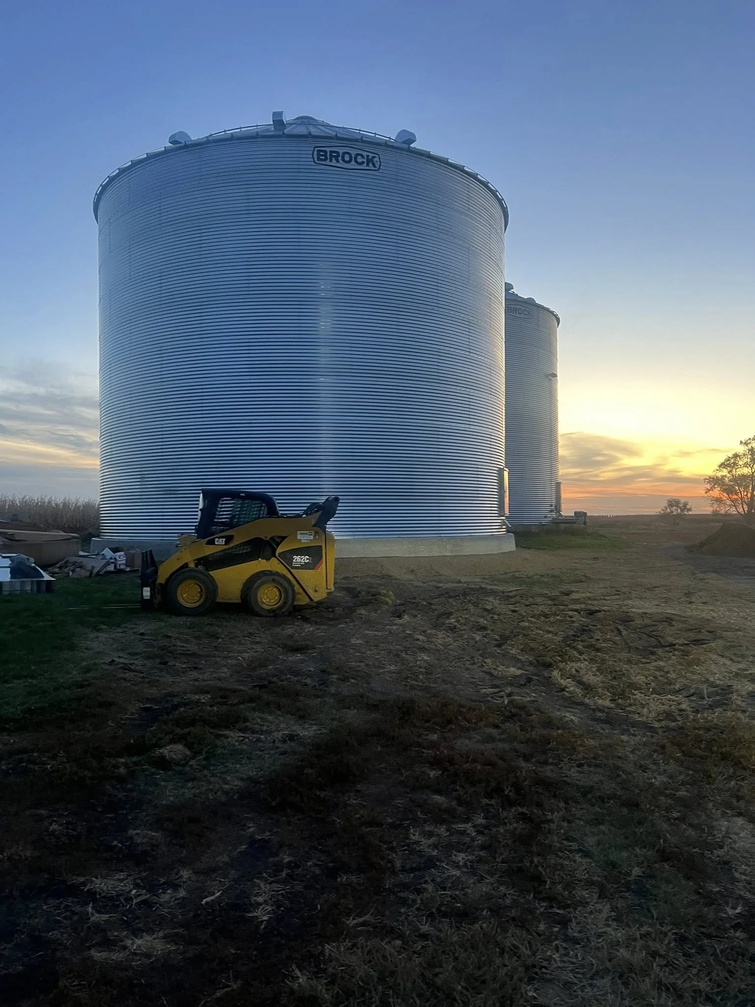 Two large silver grain silos with the word 'BROCK' on top, a yellow construction vehicle, and a sunset in the background.