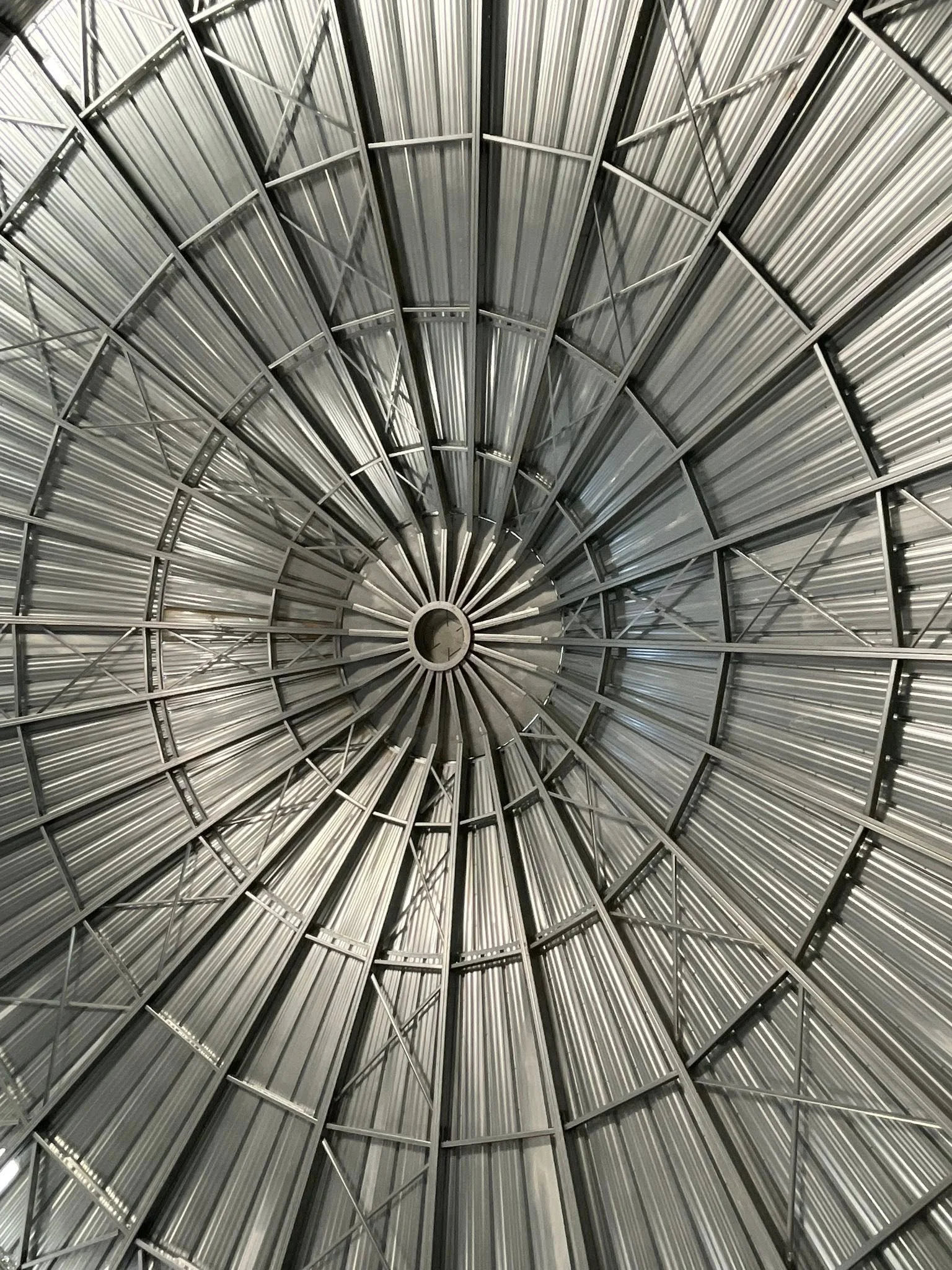 Looking up inside a circular metal structure with radial supports, resembling a dome or tunnel, made of corrugated metal panels and metal beams.