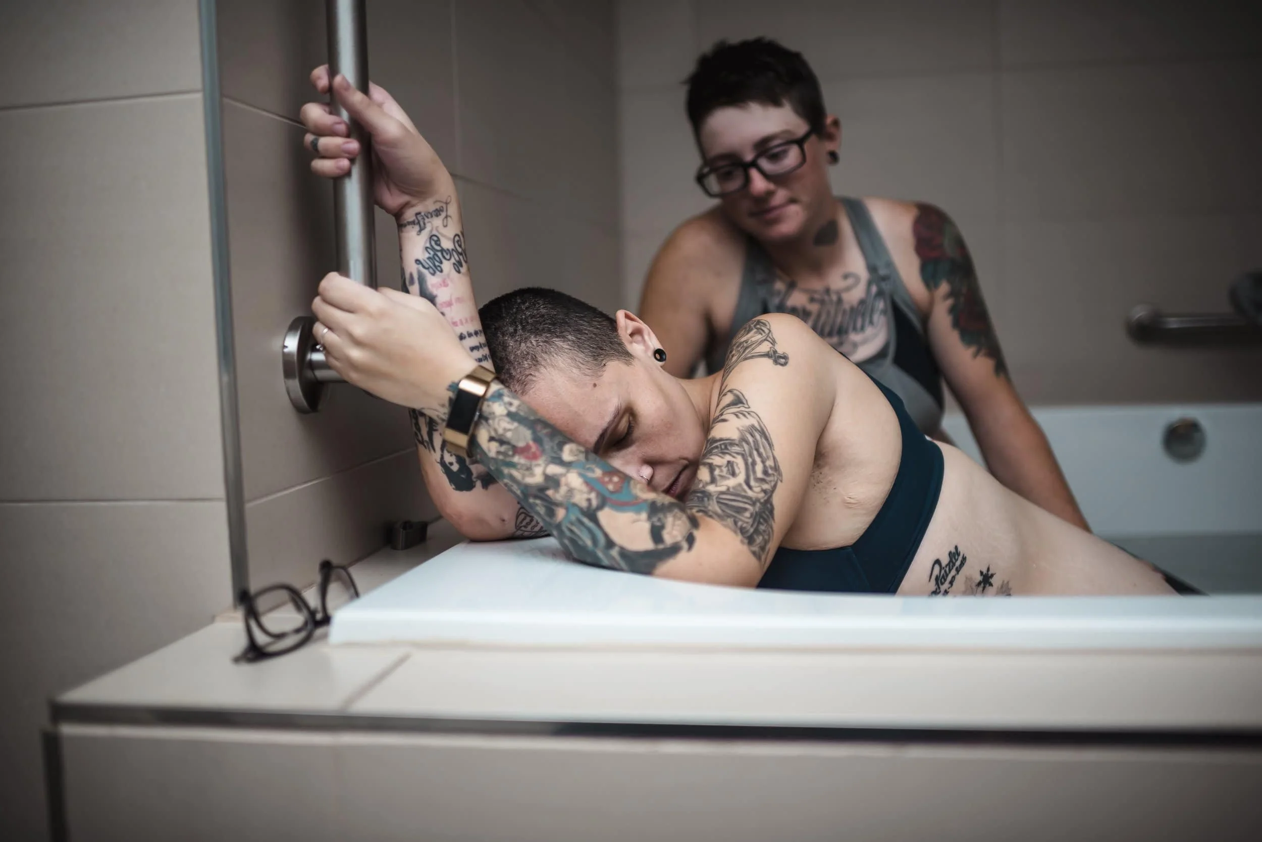 Person with beautiful tattoos holding on to support bar in the birth pool at Ottawa Birth and Wellness Centre supported by her partner.