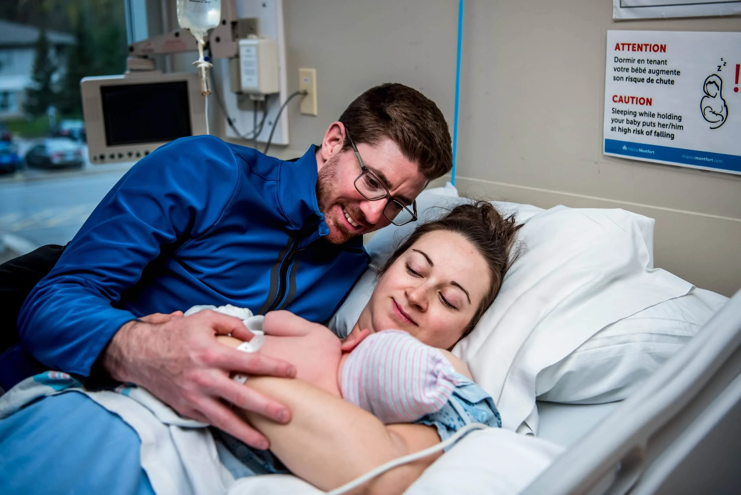 A man and a woman in a hospital room, with the woman lying in a hospital bed holding a newborn baby, and the man leaning over her, smiling. A medical monitor and an eye health warning sign are in the background.