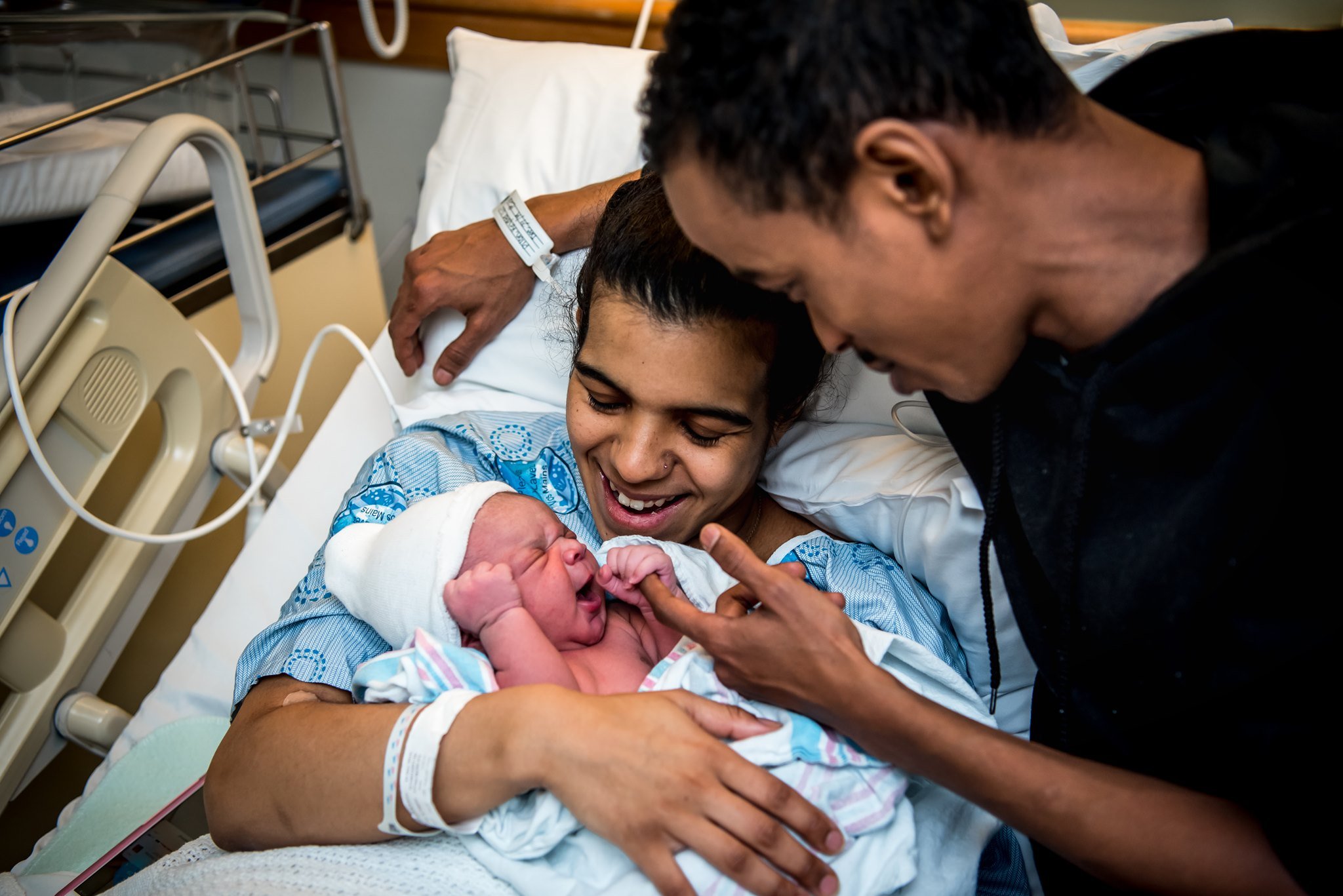 A woman in a hospital bed holding a newborn baby, with a man leaning over and touching the baby, all smiling and celebrating.