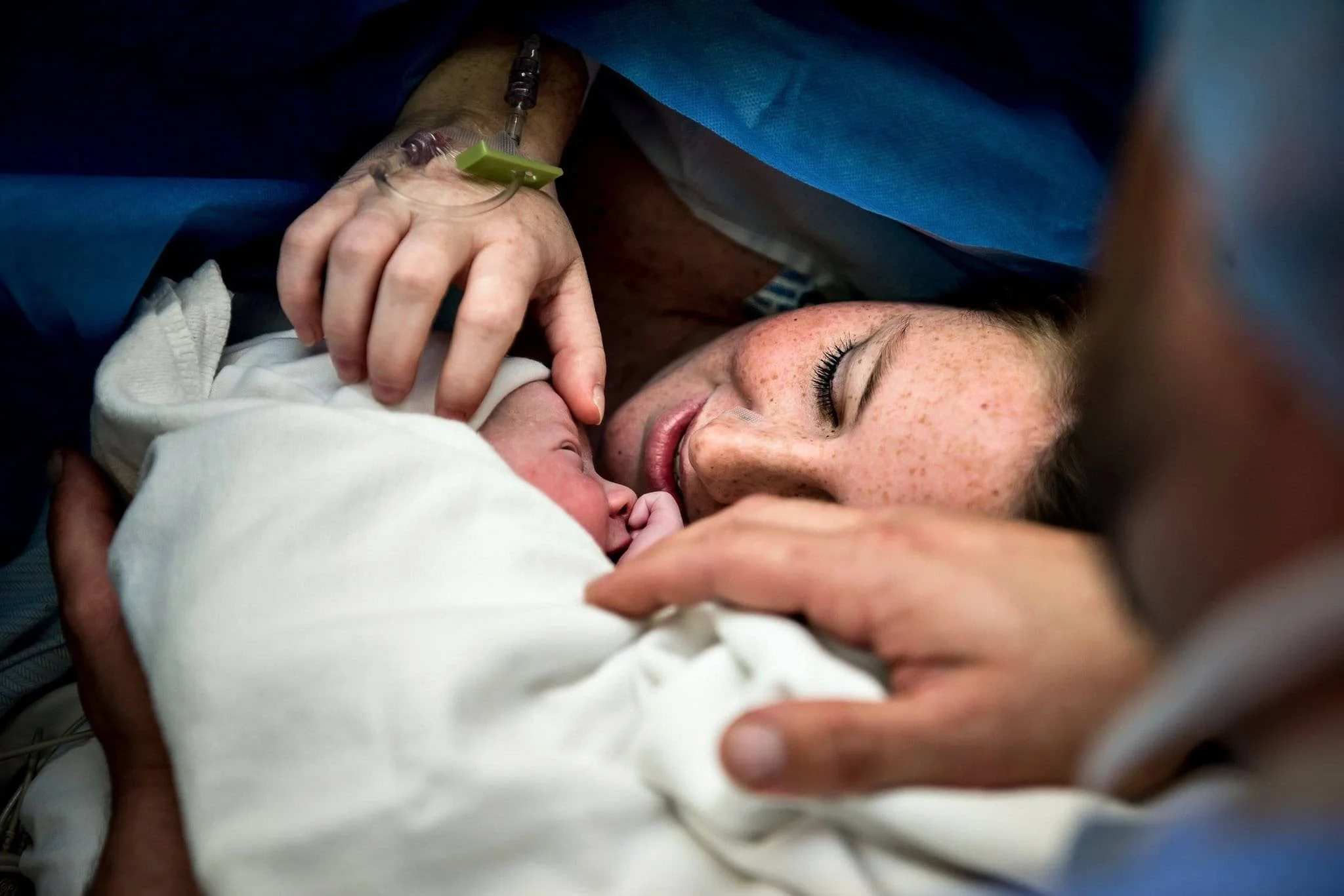 A mother and her newborn baby in a hospital, with the mother lying down and gently touching the baby, who is wrapped in a white blanket.