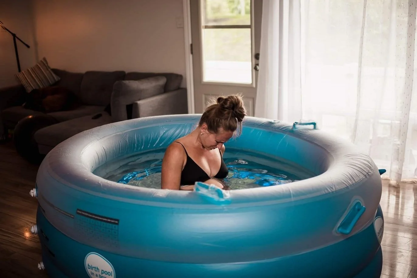 A woman in a black swimsuit sitting in an inflatable blue baby pool on a wooden floor inside a living room.
