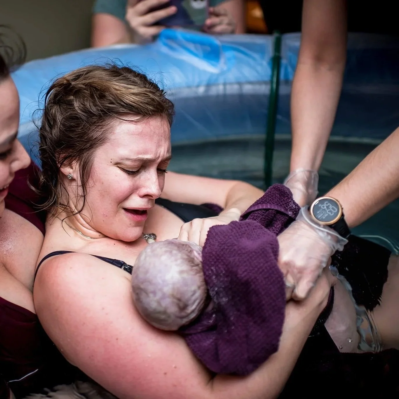 A woman giving birth in a water birthing tub, holding a newborn baby with assistance from medical personnel.