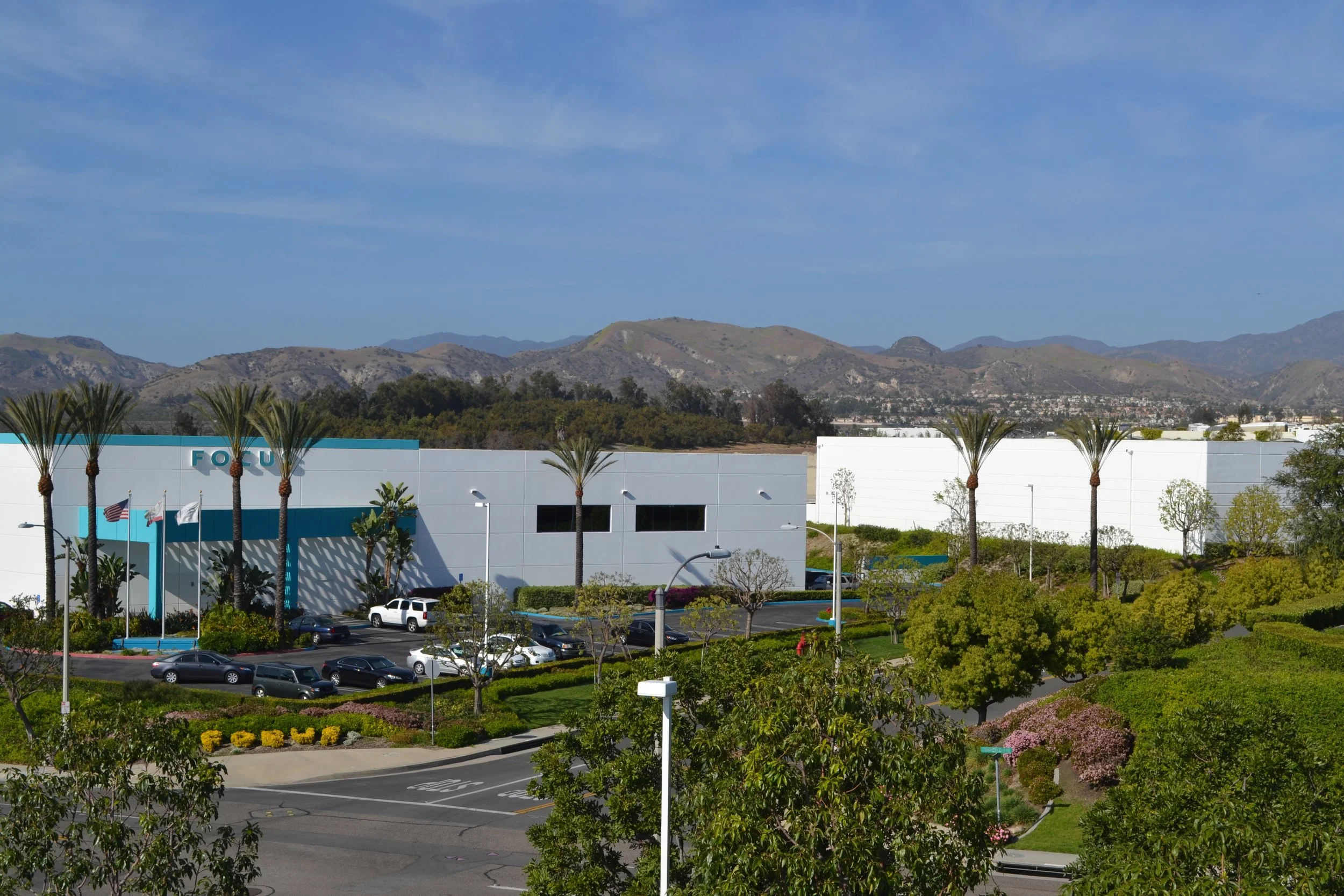 View of a shopping mall parking lot with parked cars, palm trees, and landscaping, with mountains in the background under a blue sky.