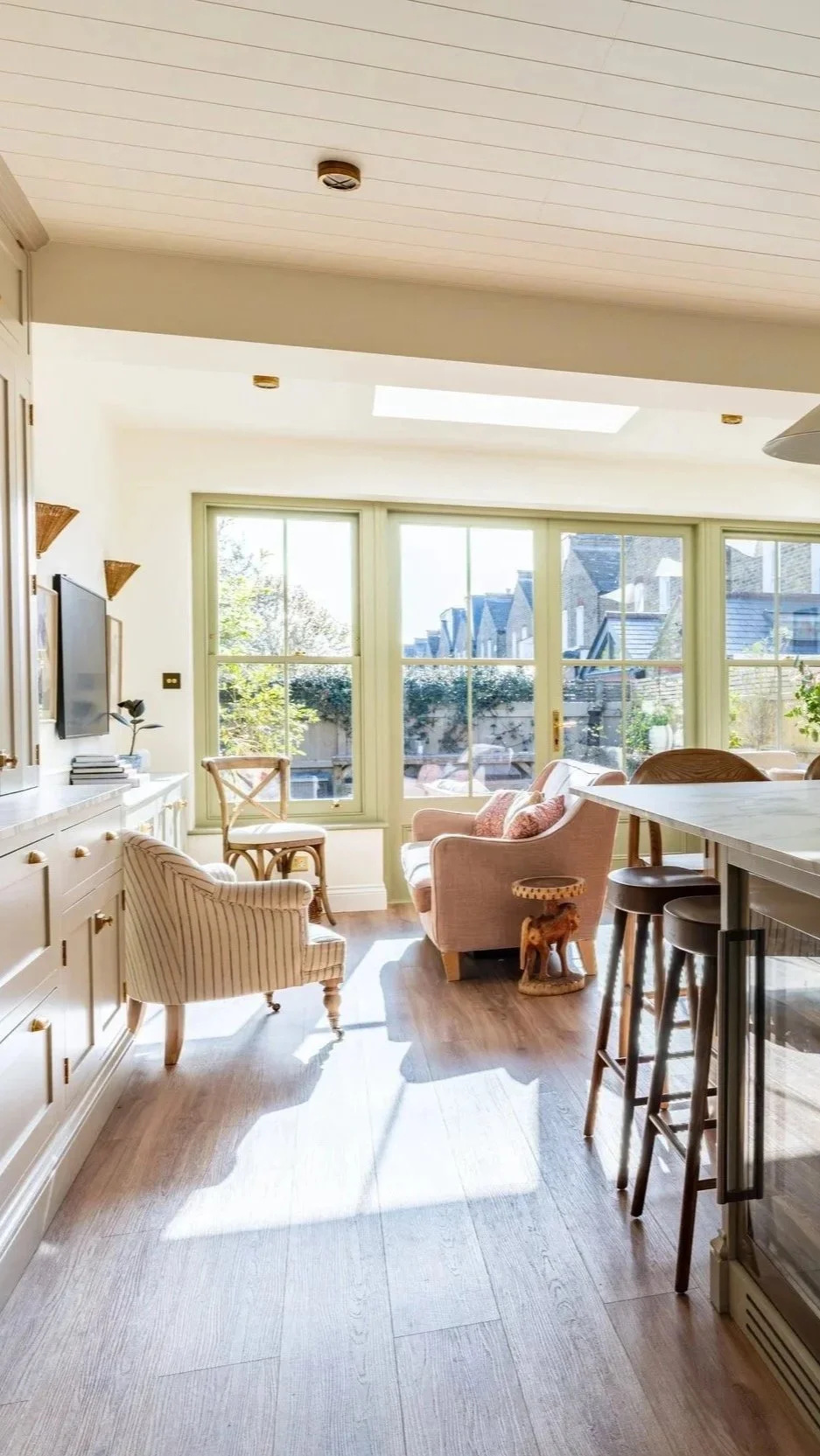 Bright living room with large windows, beige furniture, wooden flooring, and a kitchen counter with bar stools.
