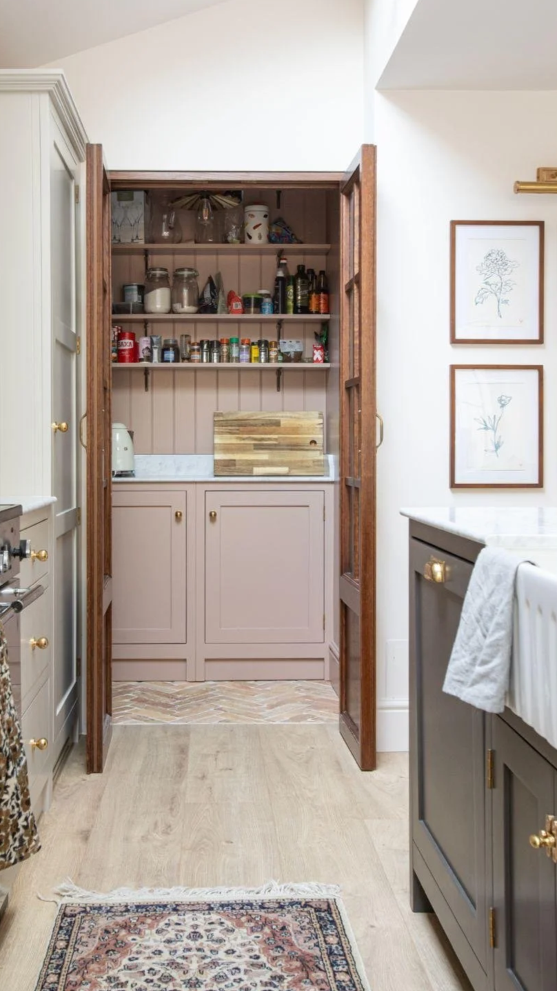 A kitchen pantry with open wooden doors revealing shelves filled with jars, spices, and kitchen items. The pantry is adjacent to a kitchen counter with a white towel hanging on a dark gray cabinet, and there are framed botanical prints on the wall to the right.