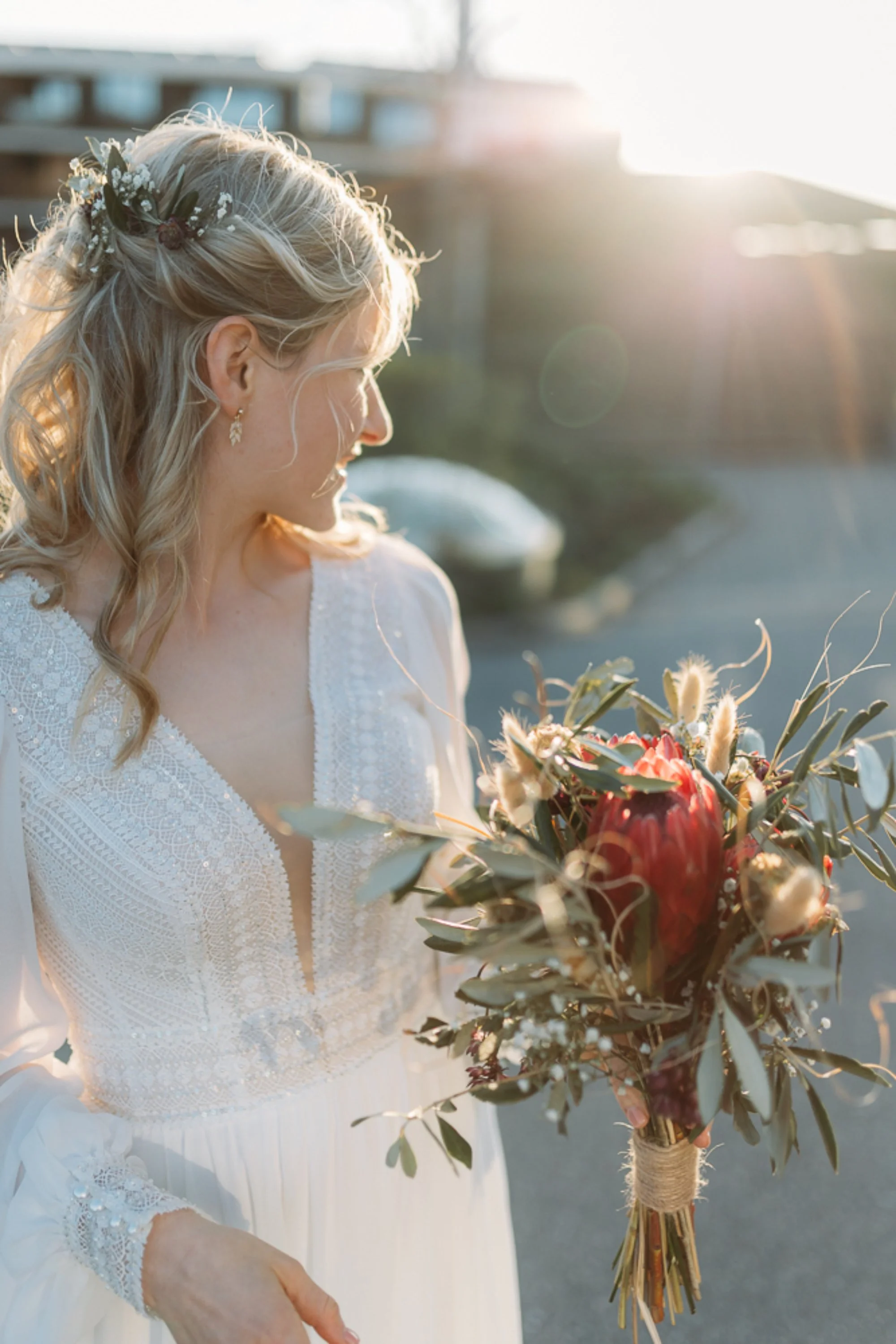 Braut in weissem Brautkleid im schönen Abendlicht, sie hält einen Brautstrauss aus roten und weissen Blumen und schaut zurück.
