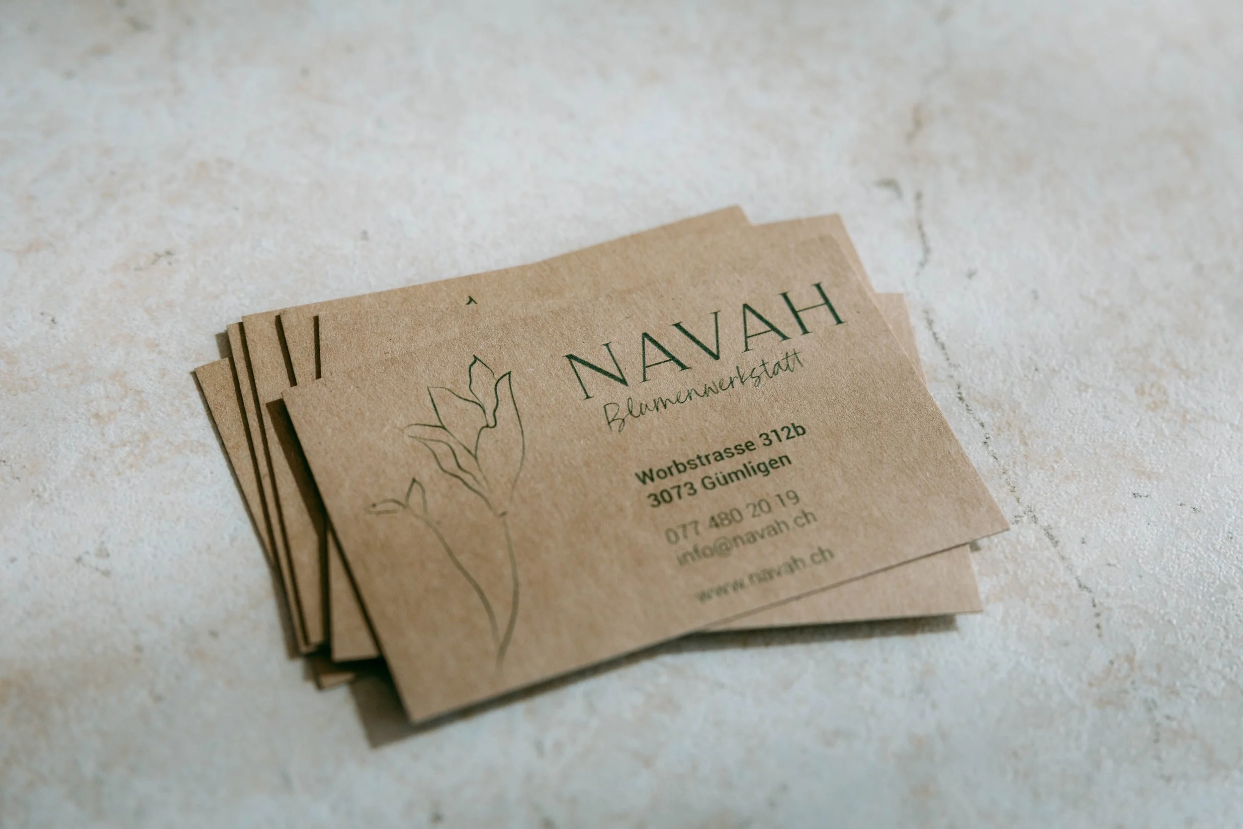 Visitenkarten für die Blumenwerkstatt Navah aus Gümlingen, mit einer stilisierten Blumenzeichnung, Kontaktinformationen und Webadresse auf braunem Papier.