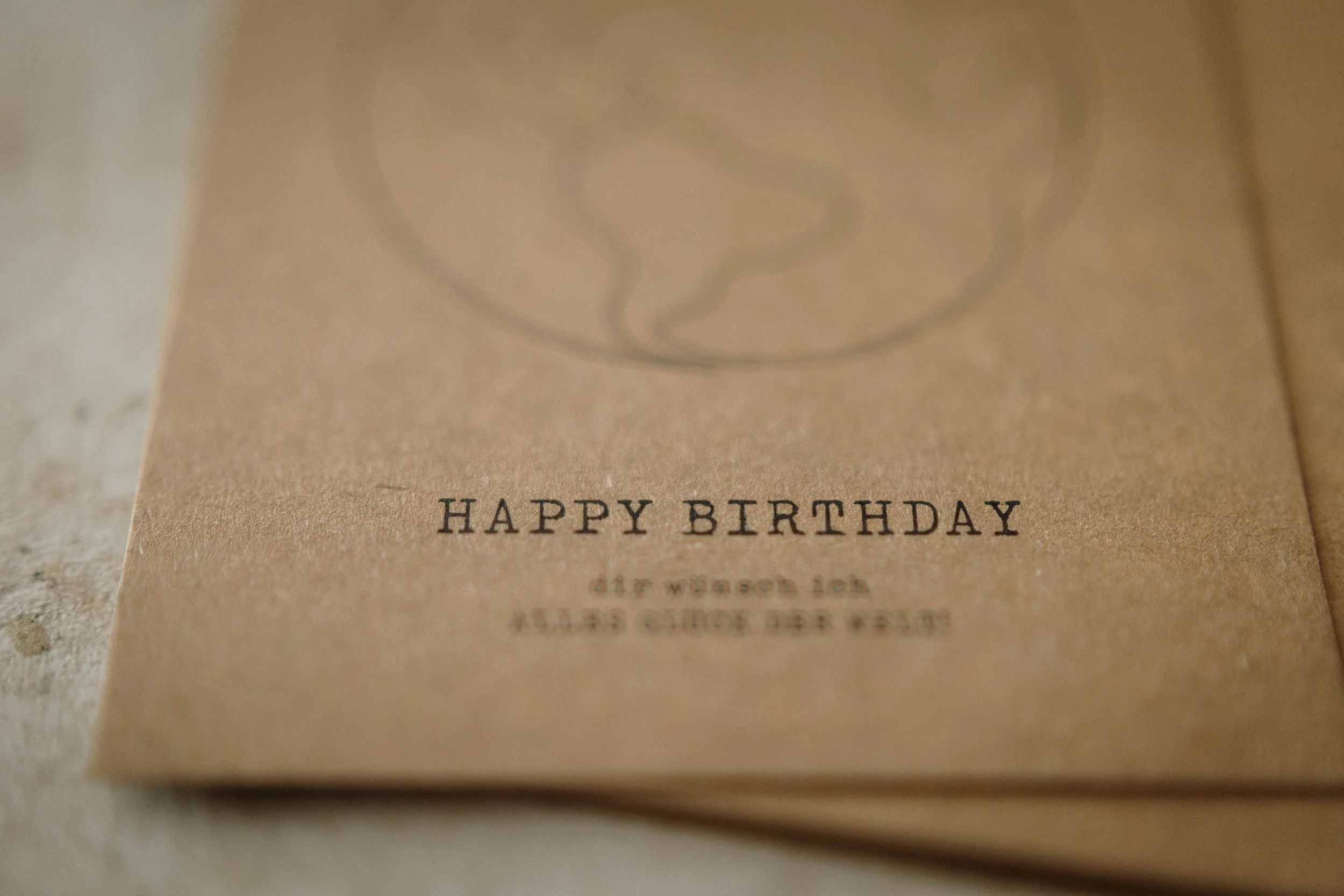 Nahaufnahme einer Postkarte mit der Aufschrift 'HAPPY BIRTHDAY' in Grossbuchstaben, auf braunem Papier, teilweise sichtbar, mit einer Zeichnung einer Weltkarte.