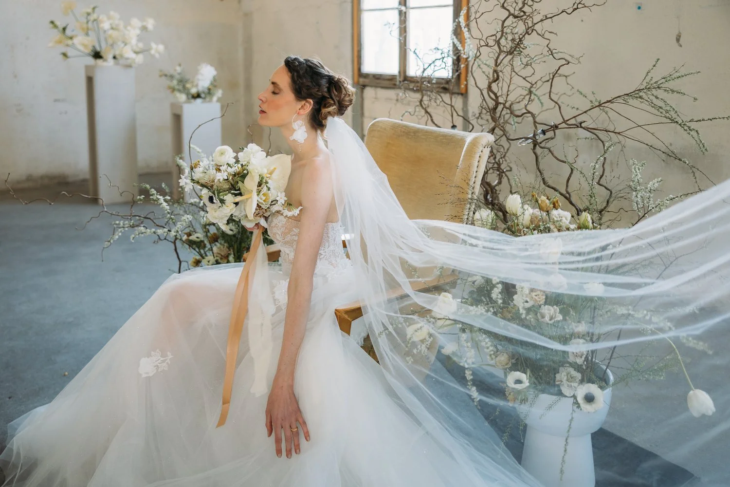 Braut in Hochzeitskleid mit Blumenstrauss, sitzend auf einer Bank bei einer Blumendekoration in einem Raum mit alten Wänden und Fenster.