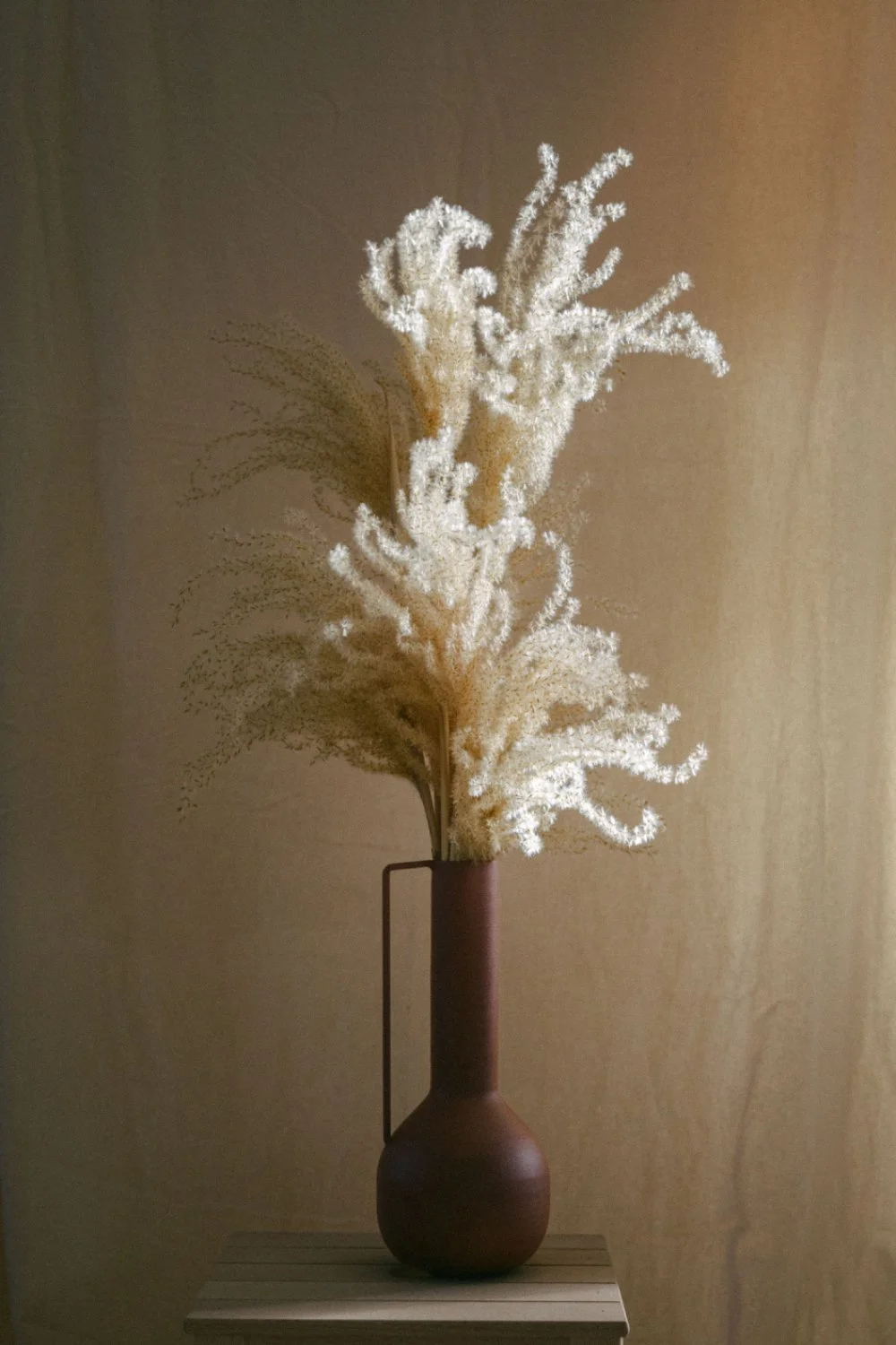 Eine braune Vase mit Pampas-Trockenblumen auf einem hellen Tischchen vor einer hellbraunen Wand.