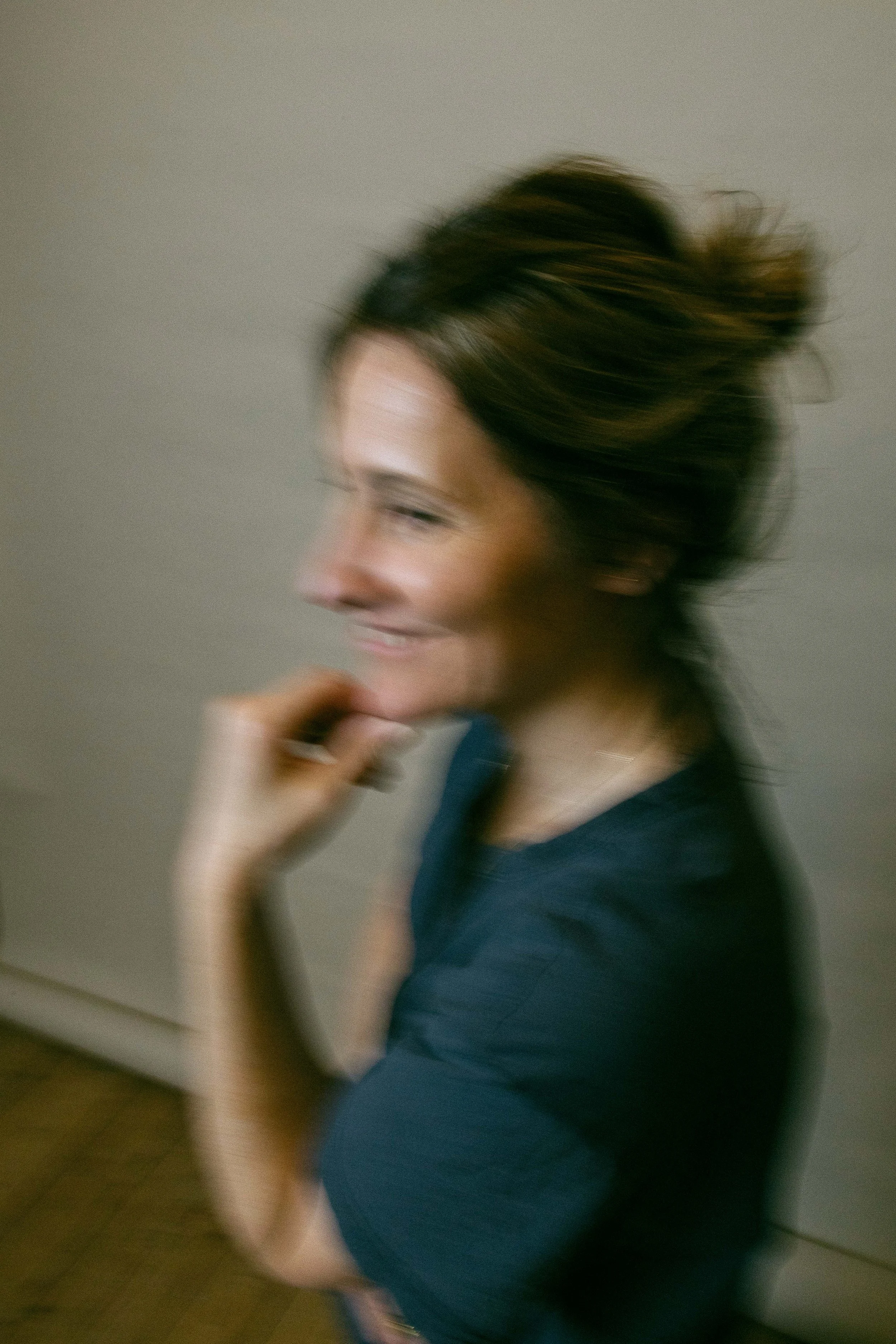 Eine lachende Frau mit braunen, gewellten Haaren, die eine Hand an den Kinn legt, auf einem grauen Hintergrund.