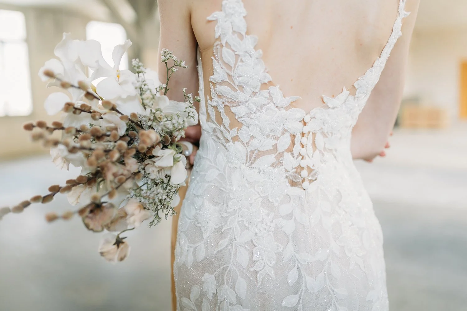 Nahaufnahme eines Brautkleides mit floraler Spitzenstickerei und einer Braut, die ein blumenreiches Bouquet hält.