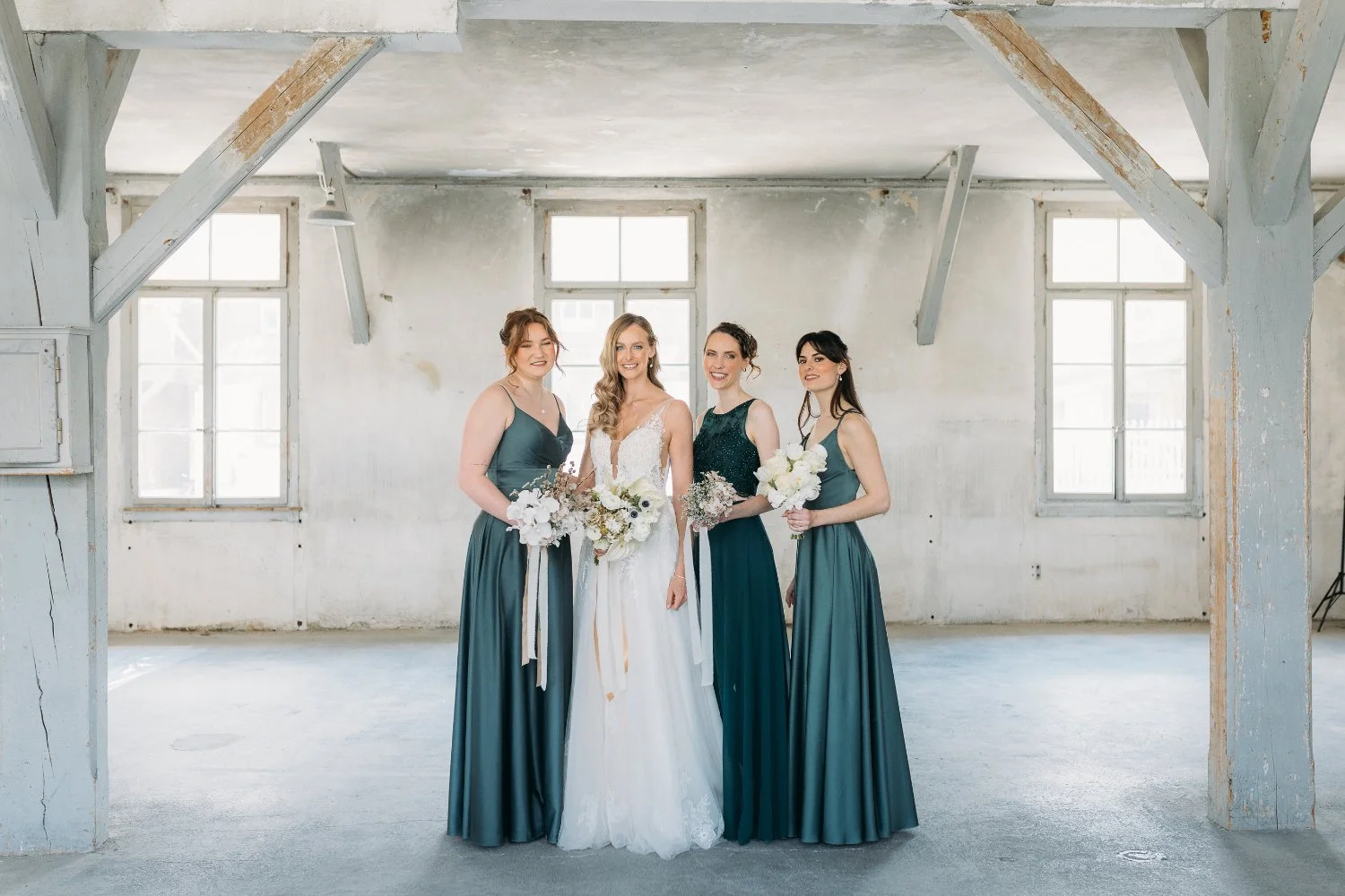 Vier Frauen, eine in einem weissen Hochzeitskleid, die anderen drei in dunkelgrünen Brautjungfernkleidern, stehen in einem hellen, industrialen Raum mit alten Fenstern, alle halten Blumensträusse.