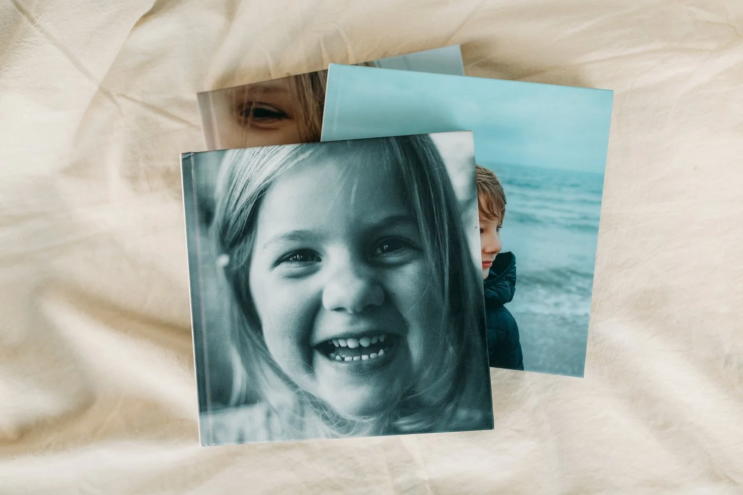 Drei Fotoalben, eines davon in Schwarz-Weiß, mit lachendem Mädchen im Vordergrund, ein weiteres mit einem Jungen am Strand im Hintergrund, auf cremefarbenem Stoff