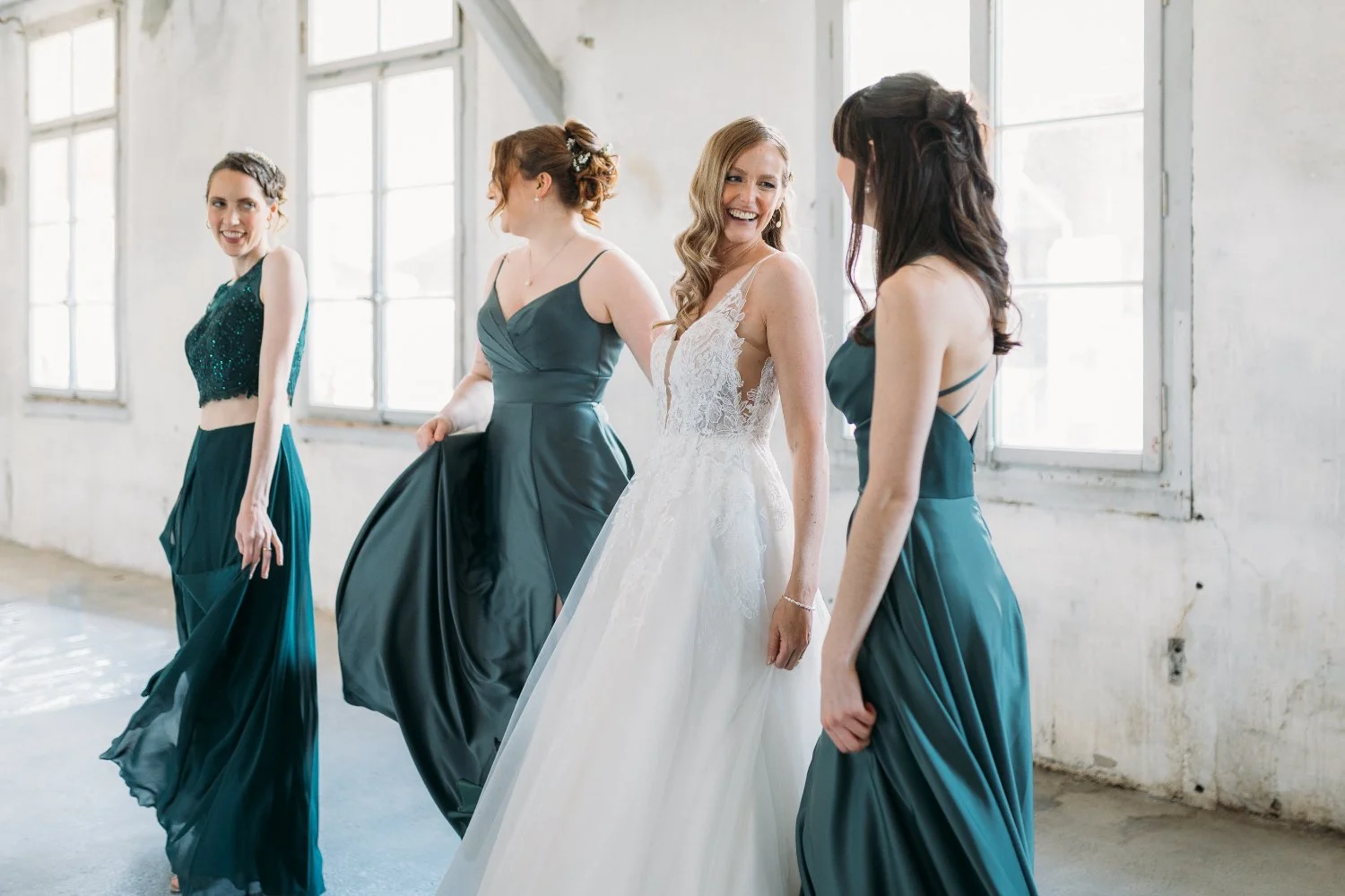 Eine Braut in einem weissen Brautkleid mit vier Freundinnen, die elegante dunkle grünfarbene Kleider tragen, in einem Raum mit grossen Fenstern und weisser Mauerwand.