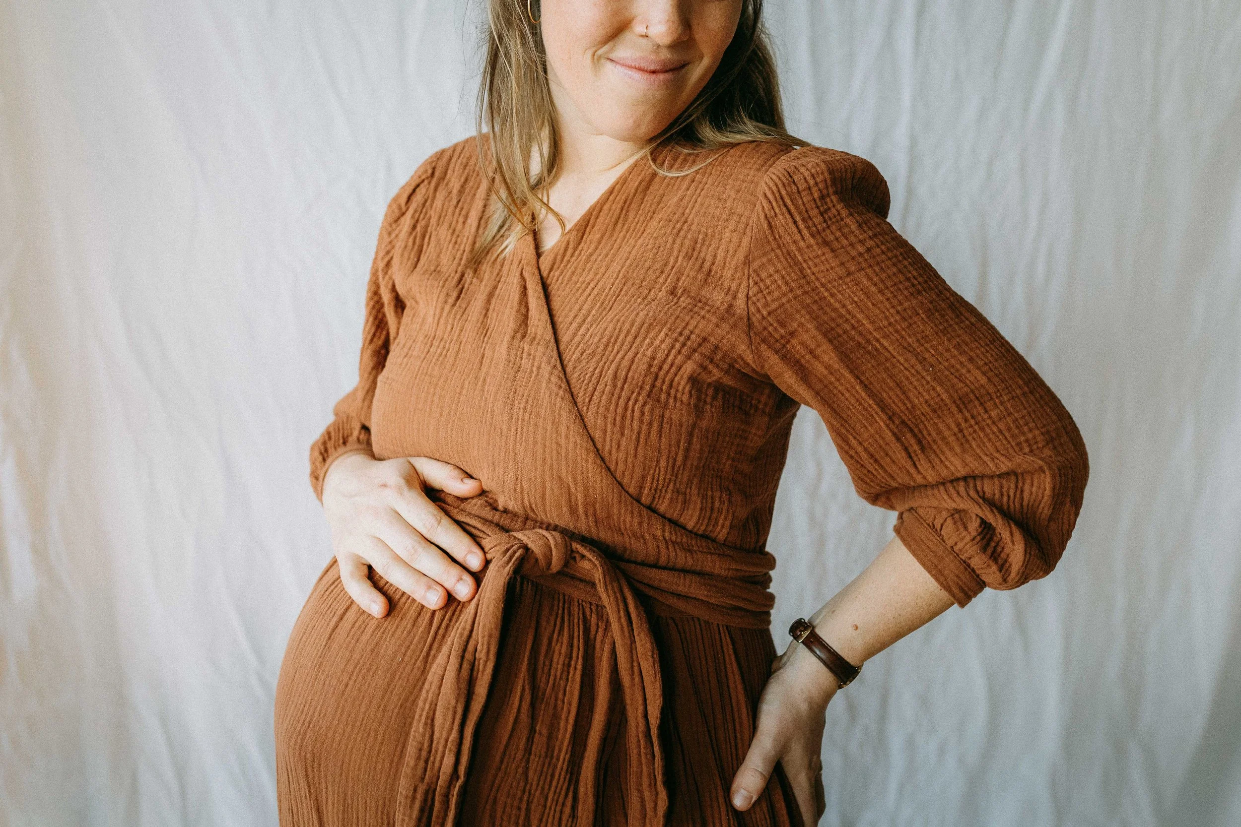 Eine Frau in einem rostroten Kleid mit halblangen Ärmeln steht vor einem weissen Hintergrund. Sie ist lächelnd und hat eine Hand an der Taille, während die andere Hand ihren Bauch hält.