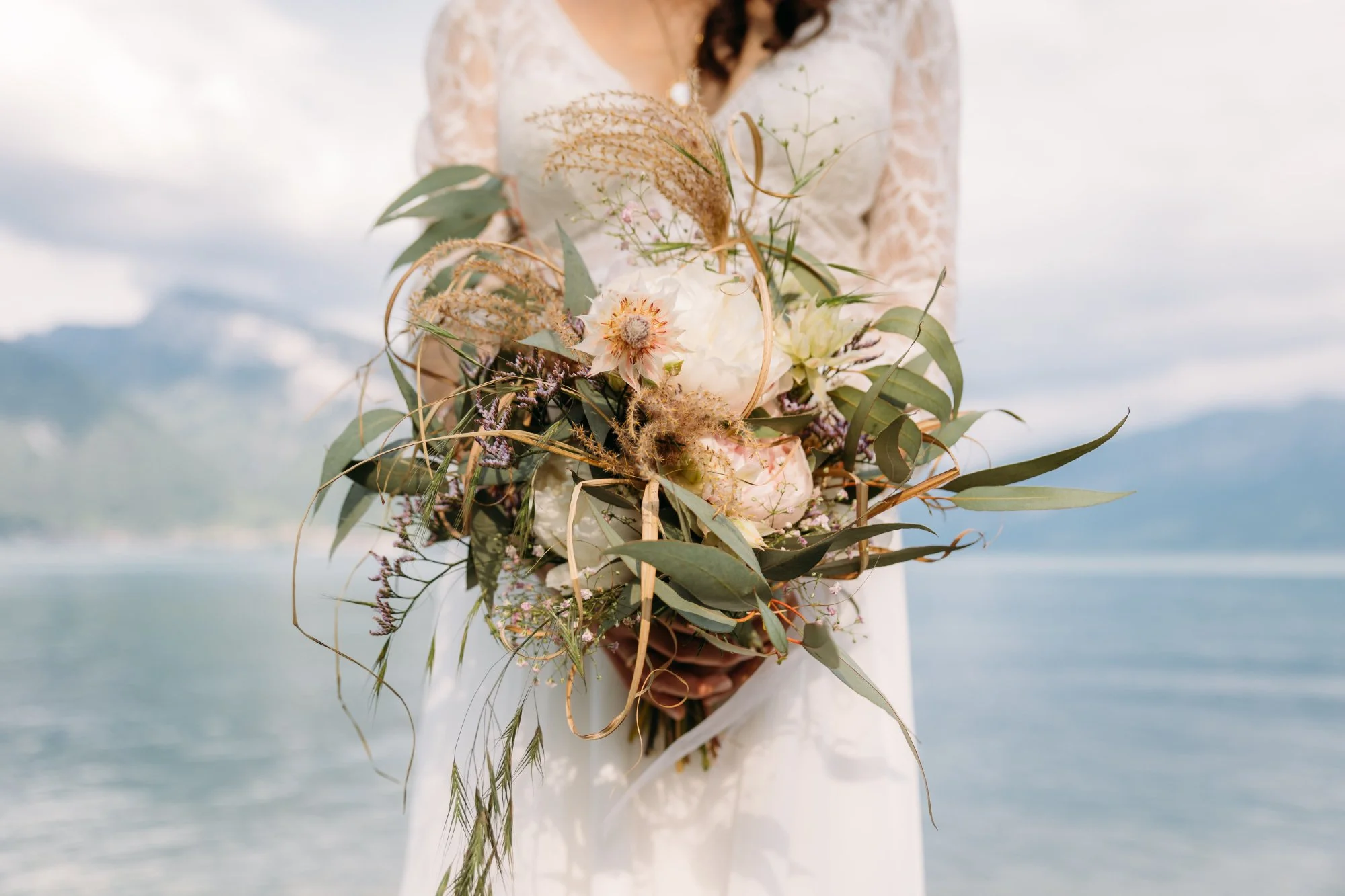 Hochzeitsbraut in weissem Kleid hält einen grossen Blumenstrauss mit weissen und rosa Blumen sowie grünen Blättern, im Hintergrund Wasser und Berge.