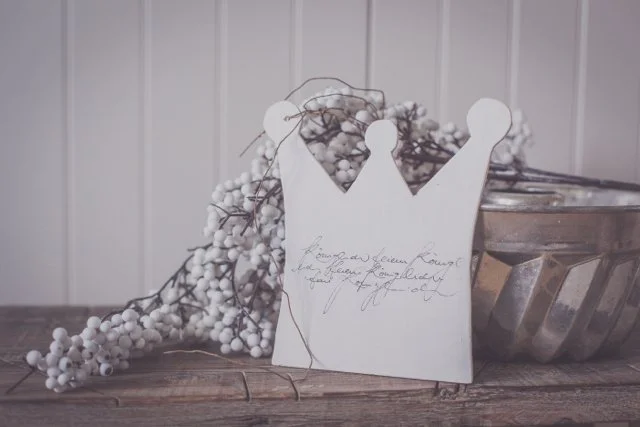 Weißes Holzkrone mit handgeschriebener Nachricht, neben weissen Beeren und Metallgefäß auf Holztisch. Hintergrund vertikale Holzpaneele.