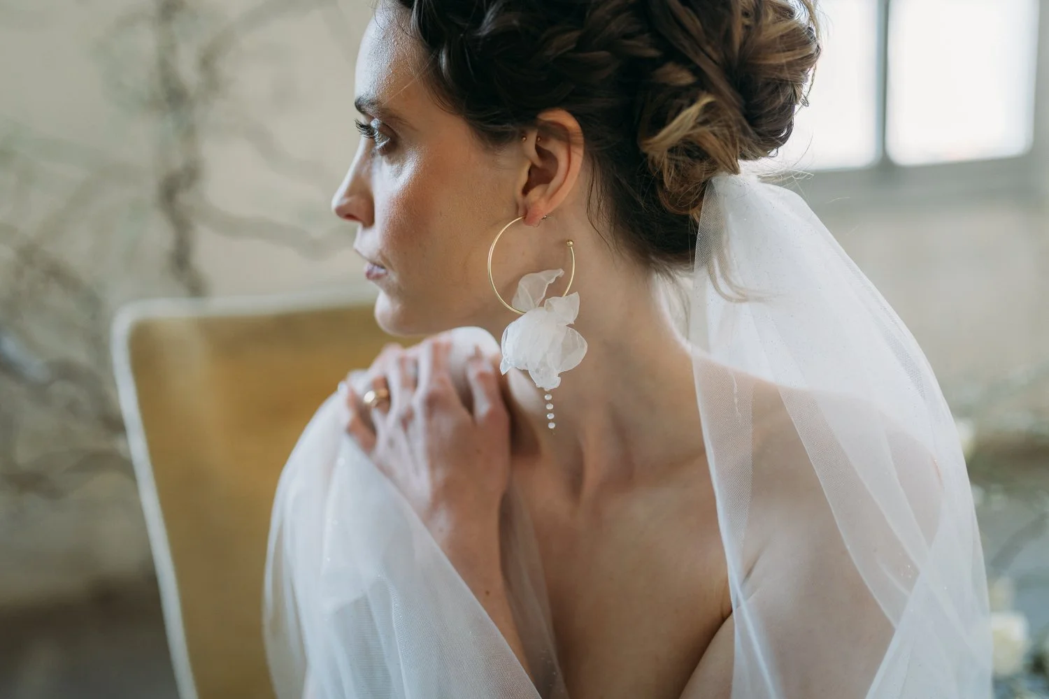 Eine Frau mit Brautkleid, grossen goldenen Ohrringen, die nachdenklich ist, sitzt in einem hell erleuchteten Raum.