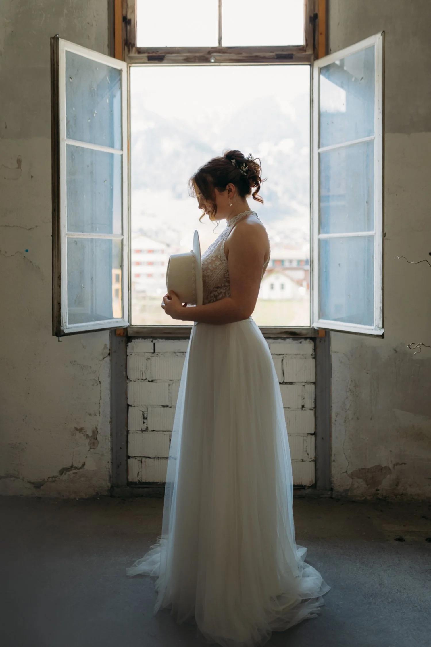 Eine Frau in einem weissen Brautkleid steht vor einem offenen Fenster, hält einen weißen Hut und schaut nach unten, während sie lächelt. Im Hintergrund sind Berge und Gebäude sichtbar.