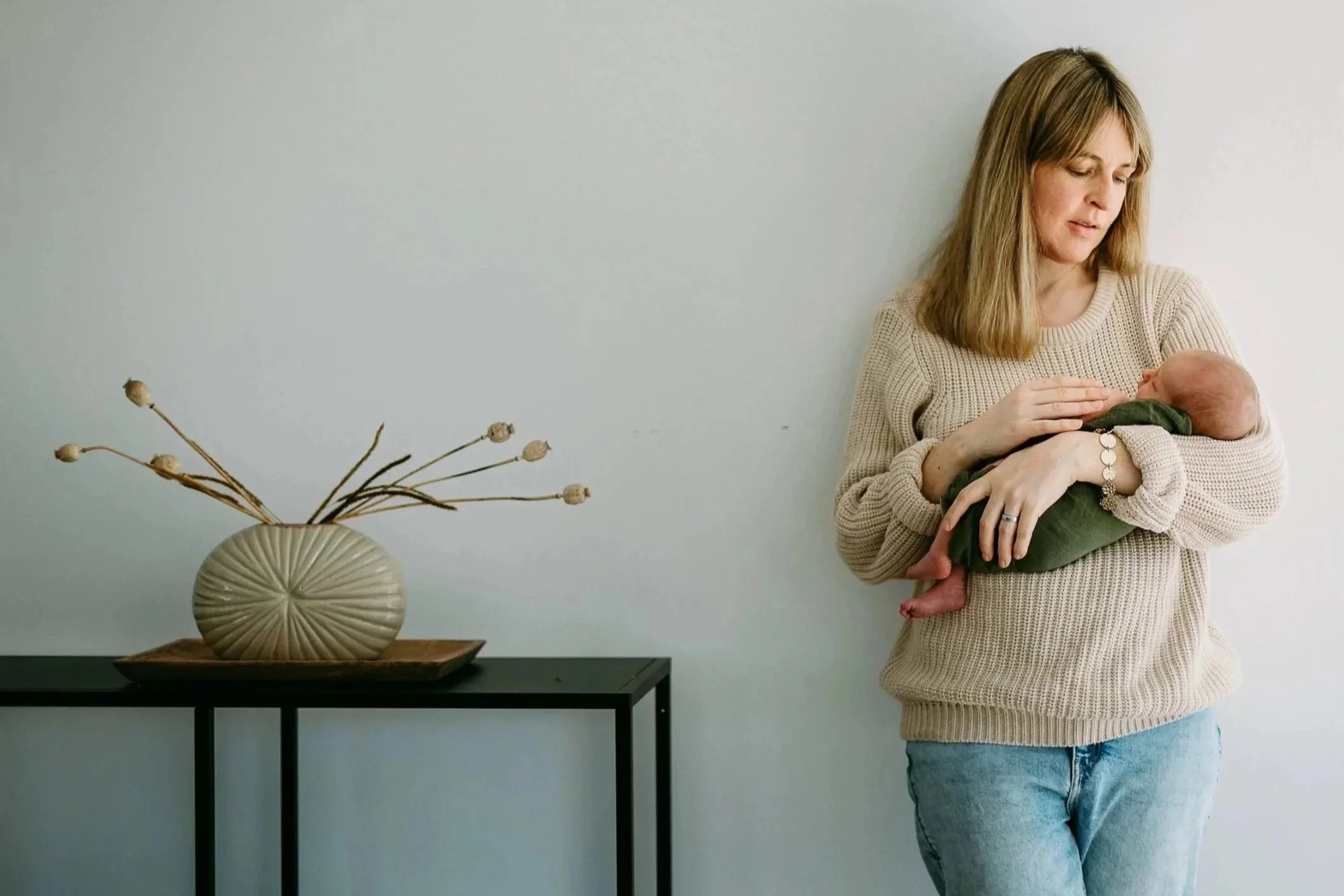 Frau hält ein neugeborenes Baby in den Armen, steht an einer weißen Wand neben einem schwarzen Tisch mit einer großen Vase. Die Frau trägt einen beige Pullover und Jeans.