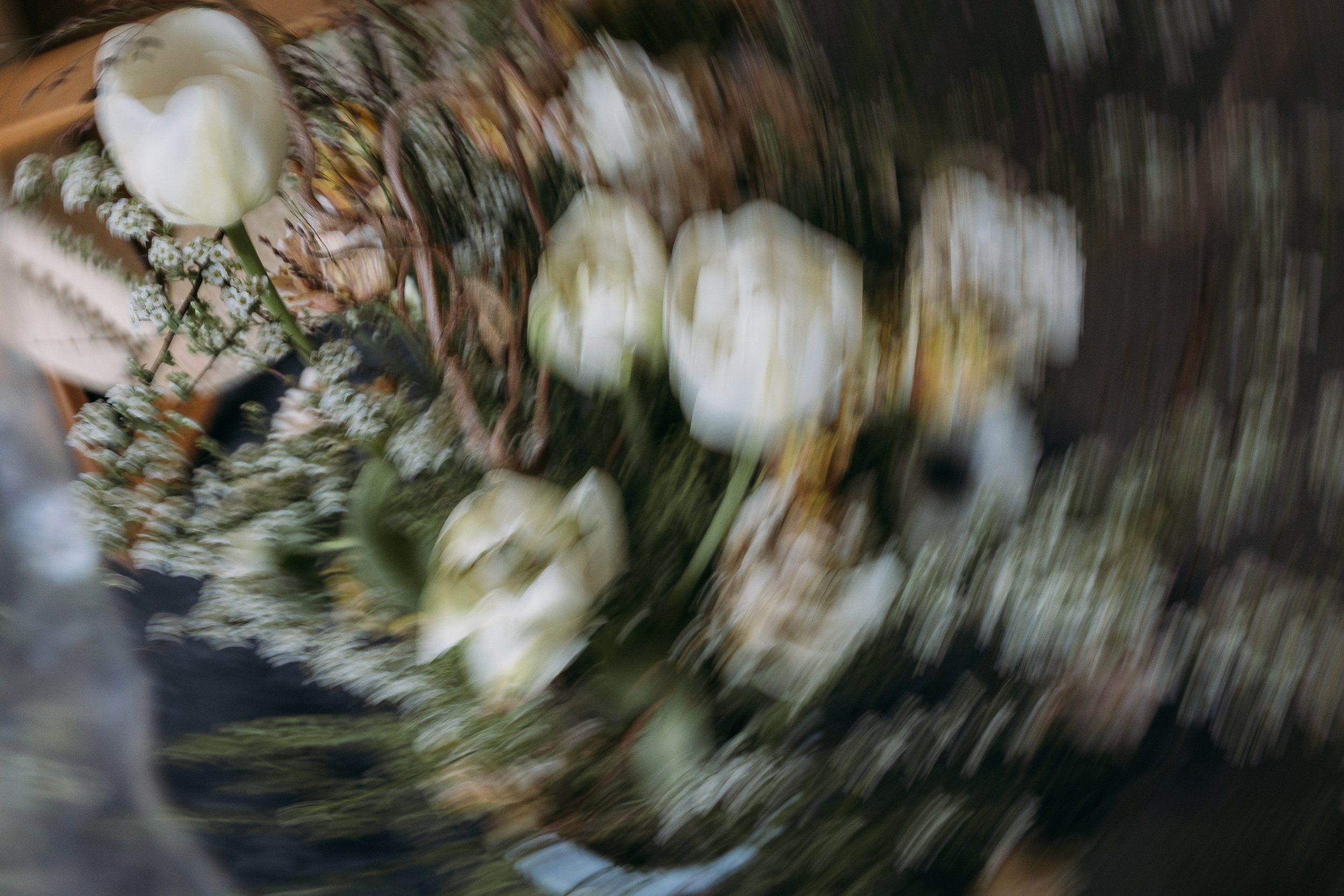 Blumengesteck mit weissen Tulpen und kleinen weissen Blumen auf einem Holztisch, unscharf und verwackelt aufgenommen.