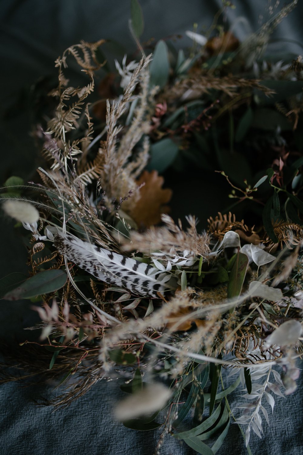 Ein dunkler Blumenstrauß mit getrockneten Pflanzen, Blättern und Feder, auf dunkelblauem Stoff.