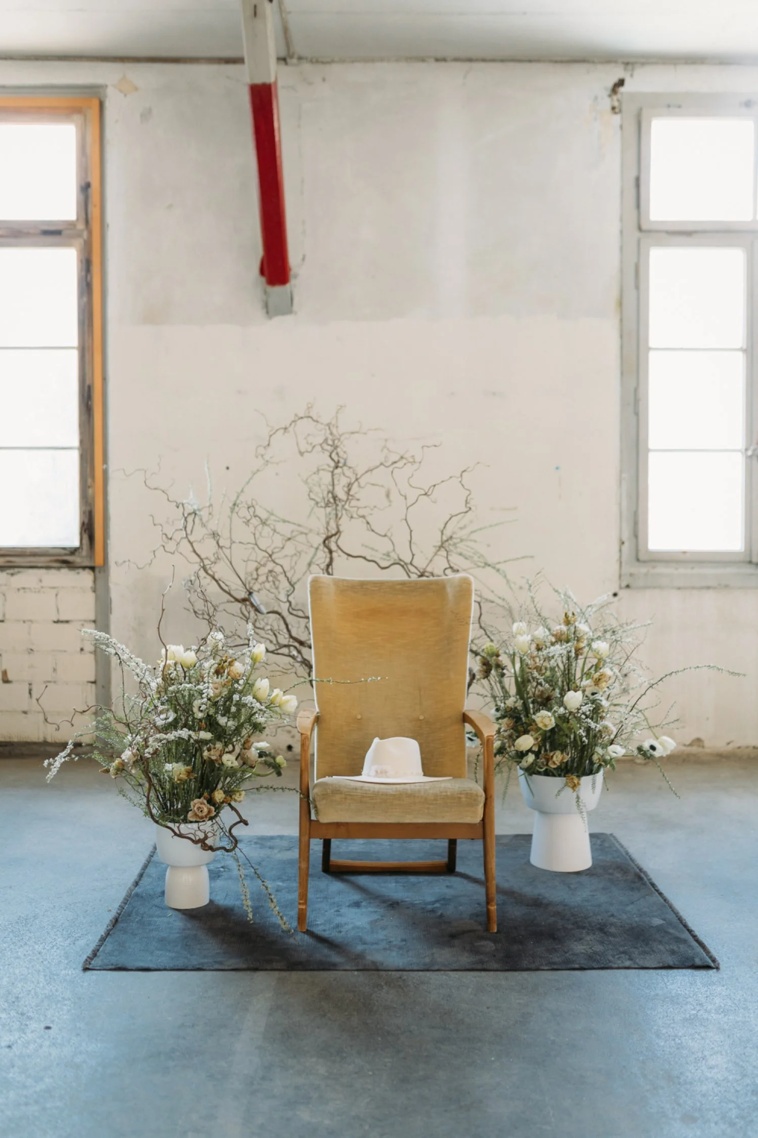 Ein stilvoller Sitzstuhl in der Mitte, umgeben von zwei weissen Vasen mit grösseren Blumensträußen, auf einem dunklen Teppich, vor einer weissen Wand mit zwei Fenstern und kunstvollen Zweigen im Hintergrund.