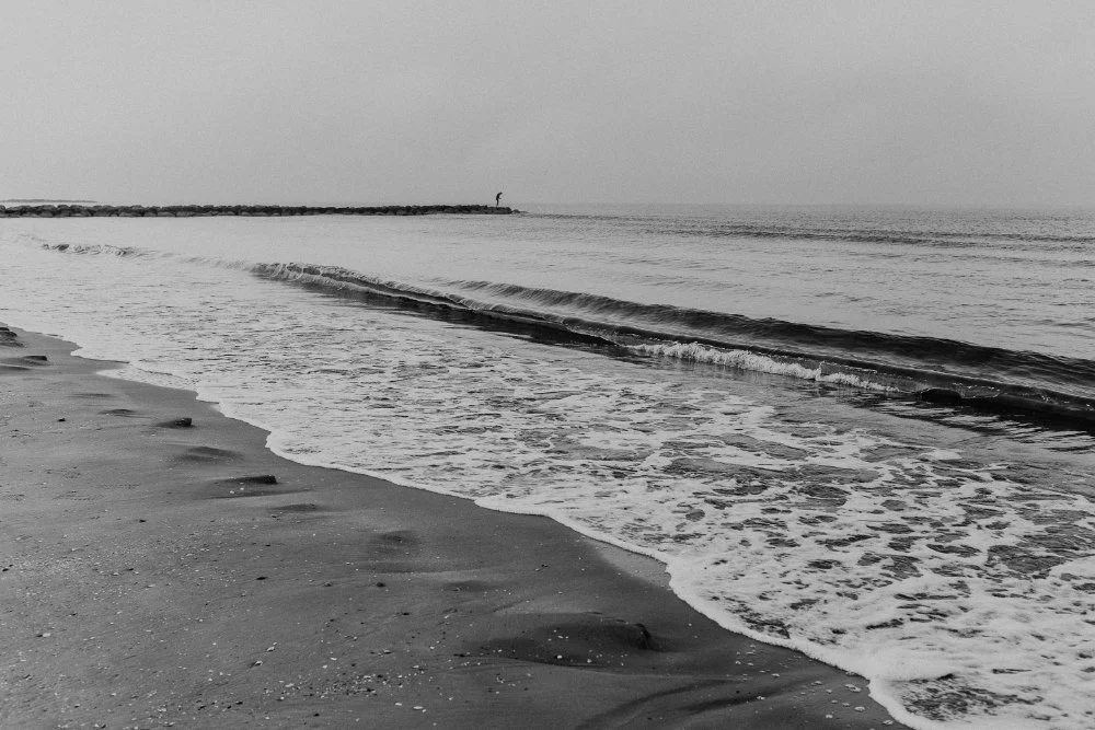 Strand mit Sand, Wellen und einem Angler in der Ferne. Schwarz-weiss-Aufnahme.
