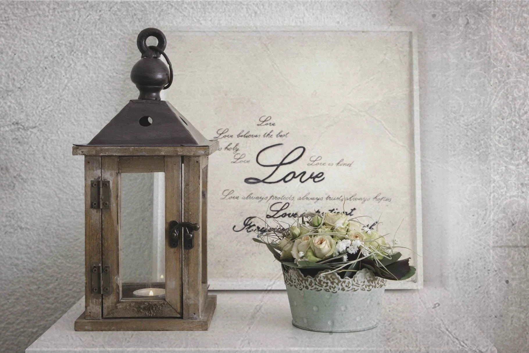 Ein Holzlaterne mit Kerze und eine Vase mit weissen Blumen auf einem Tisch vor einem Wandbild mit dem Wort 'Love' in verschiedenen Schriften.