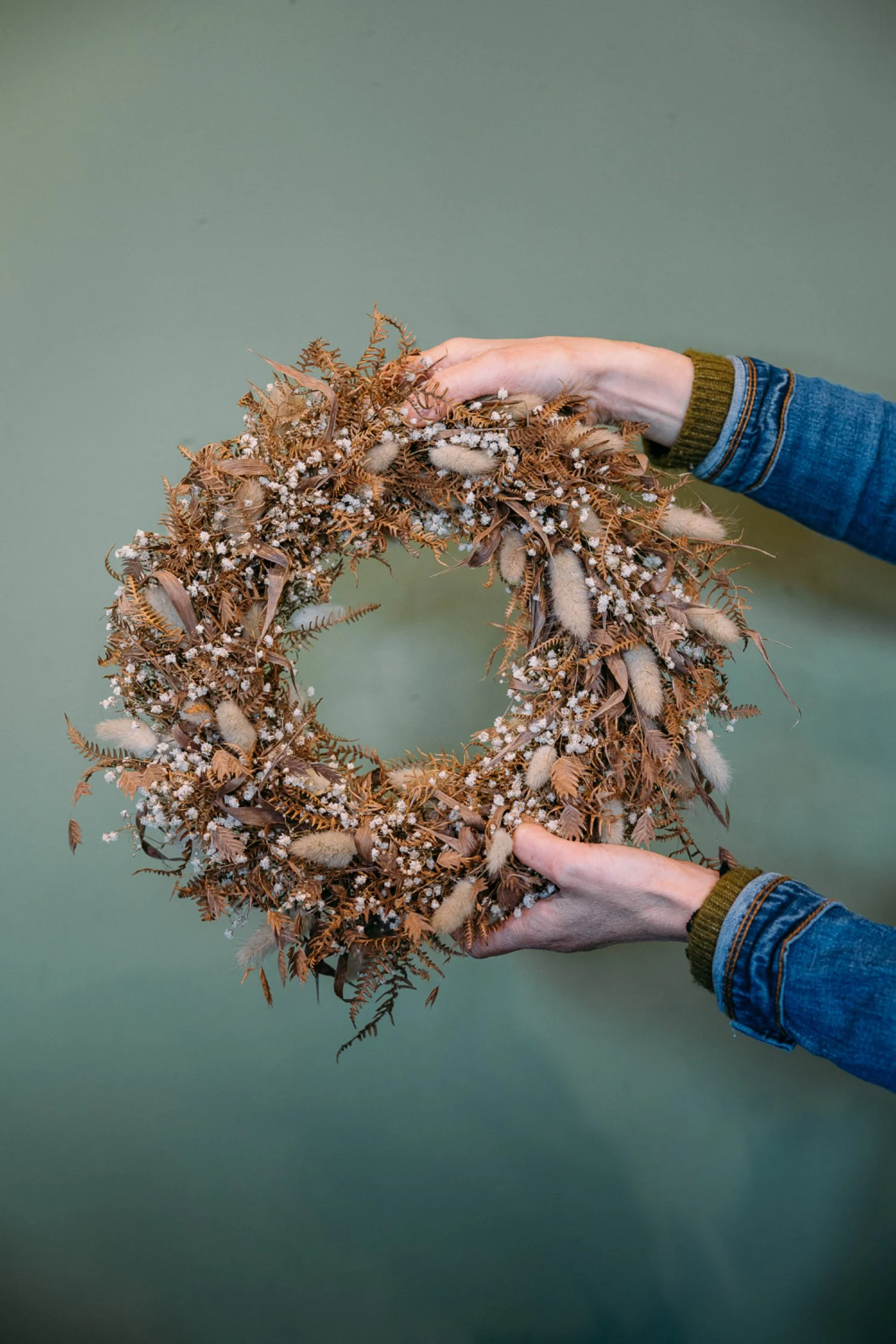 Person hält einen Herbstkranz aus getrockneten Blättern und Blumen vor einem grünen Hintergrund.