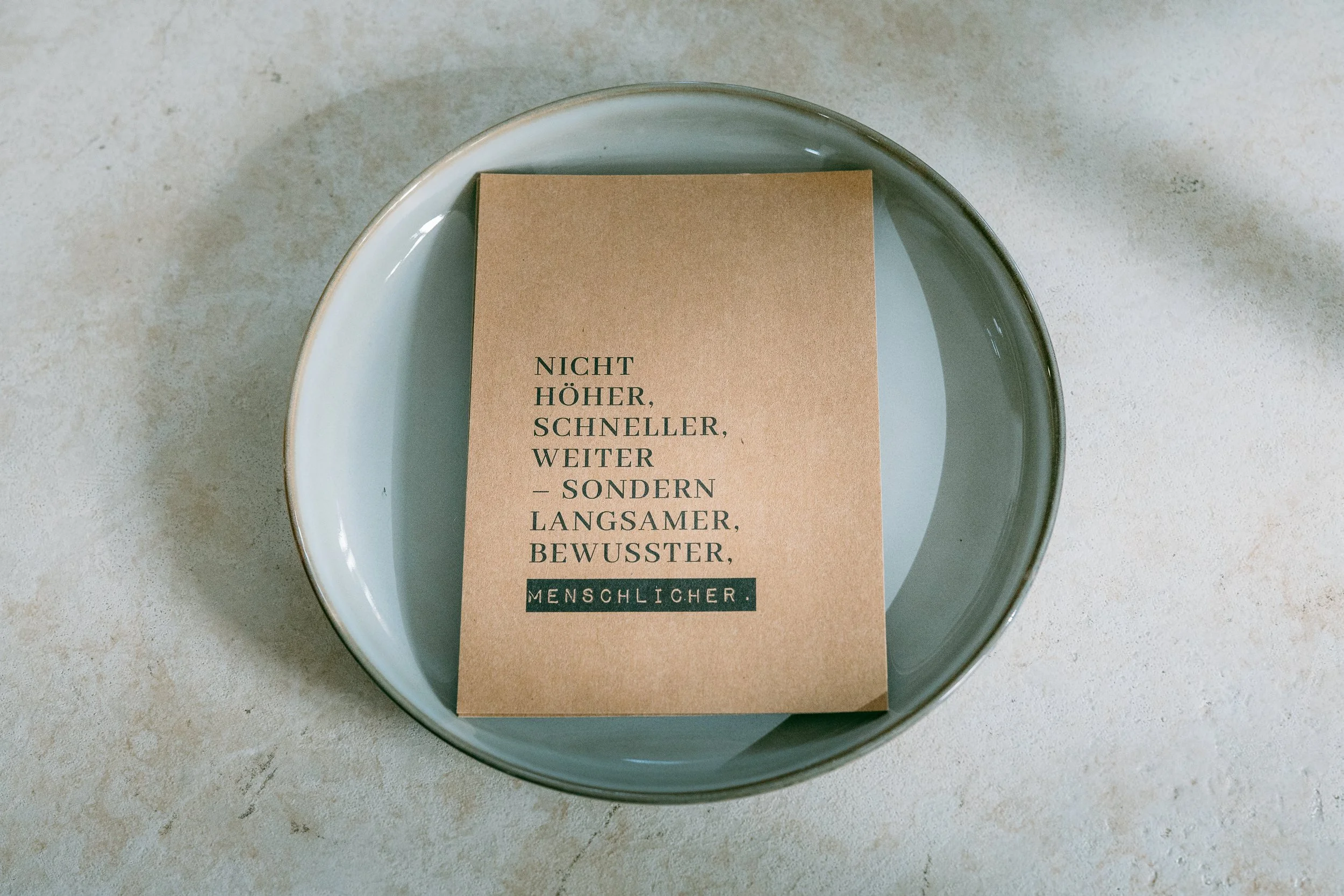 Ein braunes Papier mit einem Zitat auf einem grauen Teller. Das Zitat lautet: 'Nicht höher, schneller, weiter – sondern langsamer, bewusster, menschlicher.'