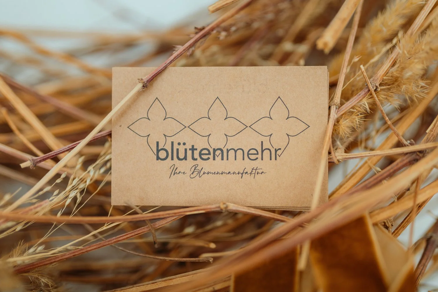 Ansicht einer braunen Visitenkarten mit dem Logo "blütenmehr" und dem Schriftzug "Ihre Blumenmanufaktur", umgeben von getrockneten Pflanzen und Zweigen.