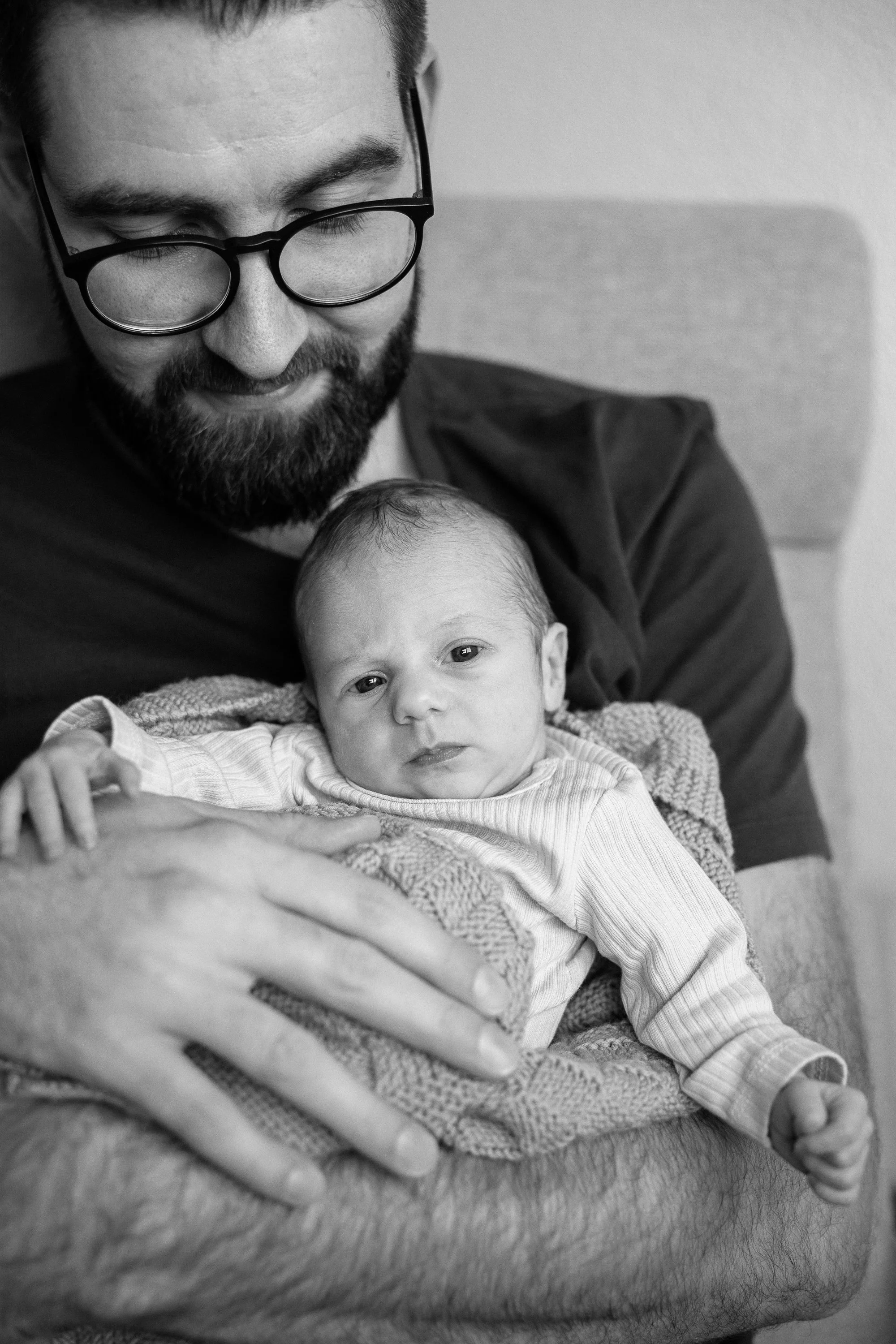 Ein Mann mit Brille hält ein Baby auf seinem Arm, beide sitzen auf einem Sofa. Das Baby schaut direkt in die Kamera, während der Mann lächelt. Das Bild ist schwarz-weiss.