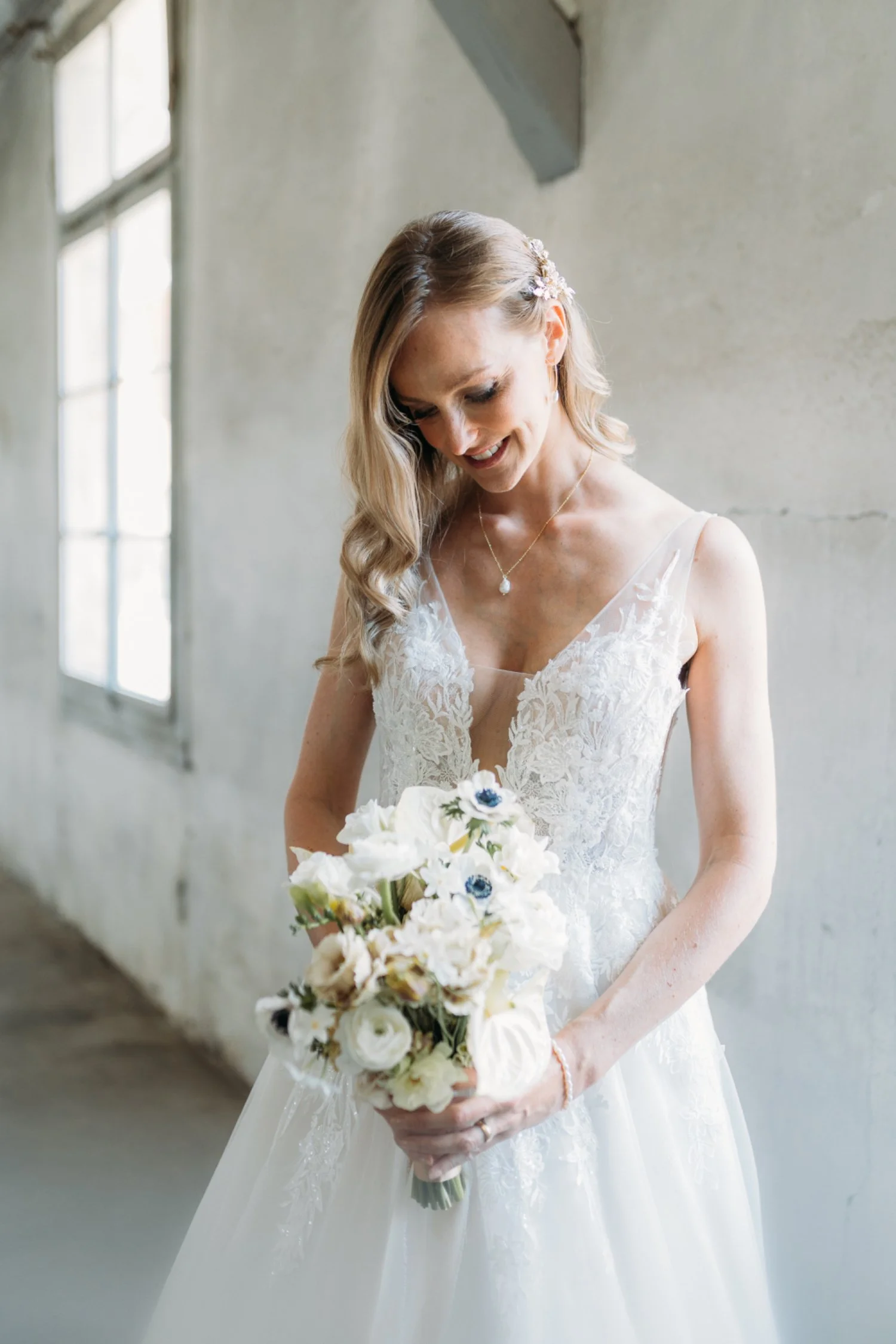 Braut in weissem Hochzeitskleid hält einen Blumenstrauss und blickt nach unten, Hintergrund mit grossen Fenstern in einem hellen Raum.