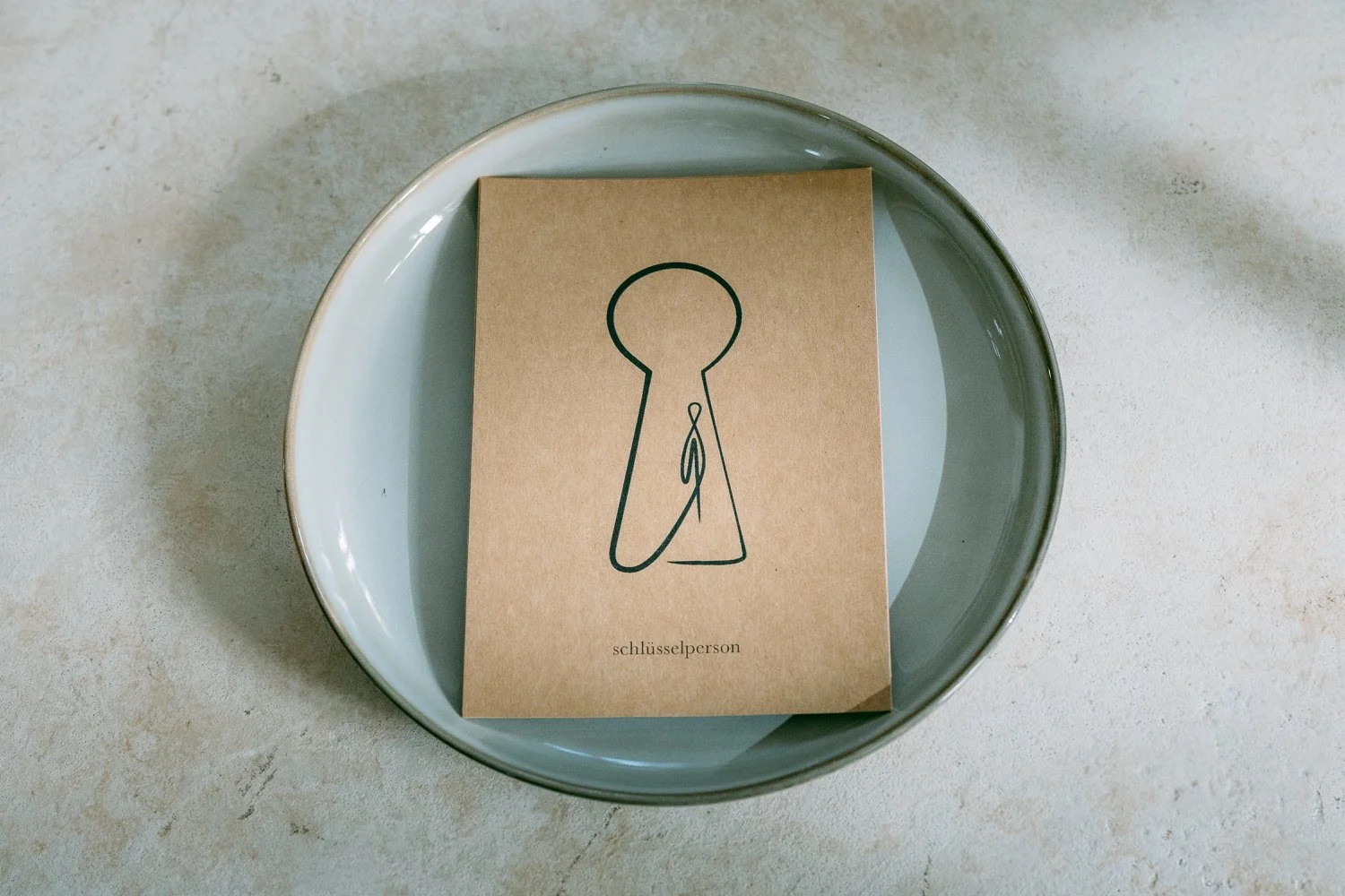 Karte mit einer minimalistischen Illustration eines Schlüsselperson-Symbols, auf einem grauen Teller liegend, mit dem Wort 'schlüsselperson' am unteren Rand.