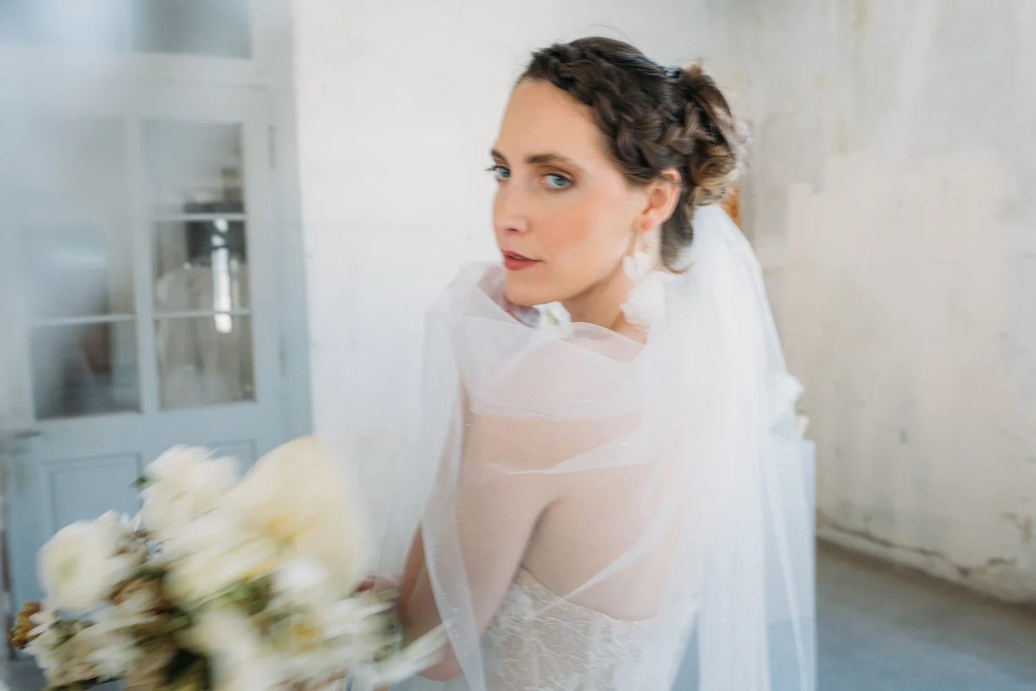 Frau in Hochzeitskleid mit Blumenstrauss, blickt in Kamera, in heller, minimalistischer Umgebung.