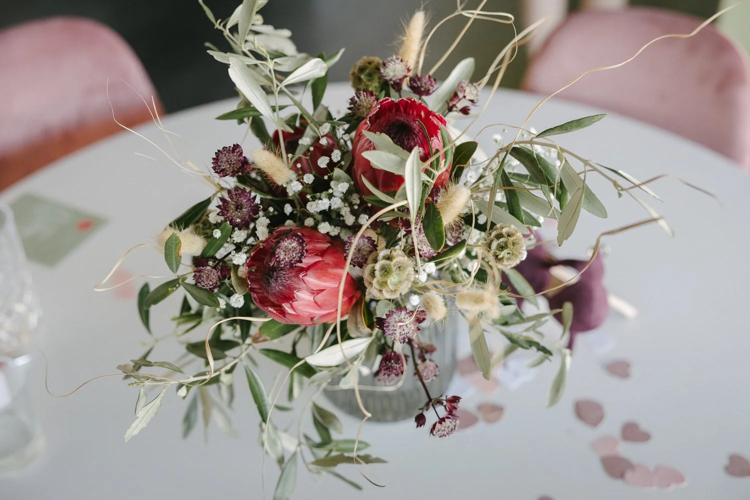 Blumenstrauß mit roten Protea, weißen, lila und grünen Blumen und Blättern auf einem weissen Tisch, umgeben von rosa Herzchen.