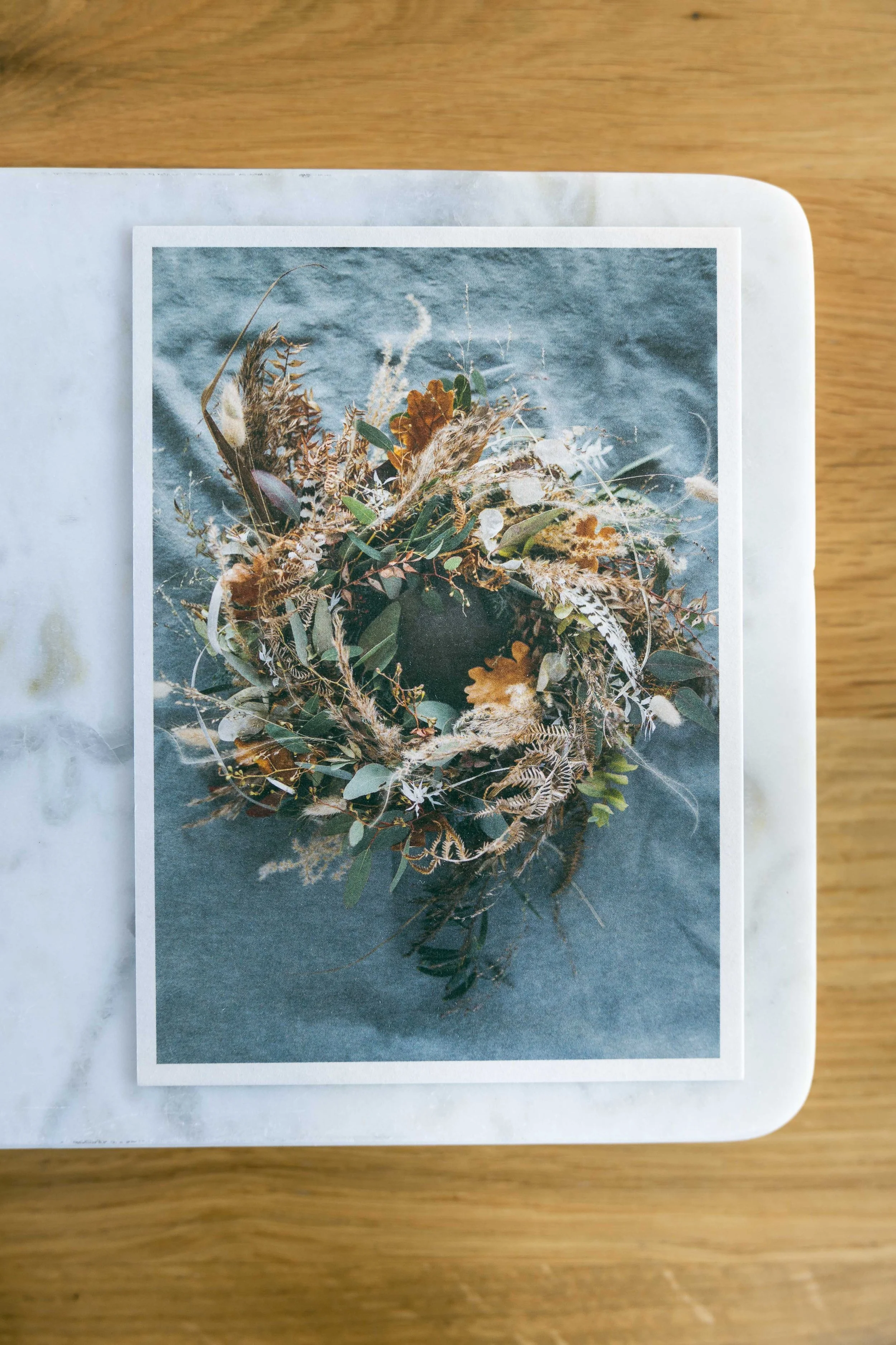 Fotografie eines Blumenkranzes mit verschiedenen getrockneten Gräsern und Blättern, auf einem grauen Stoffhintergrund.