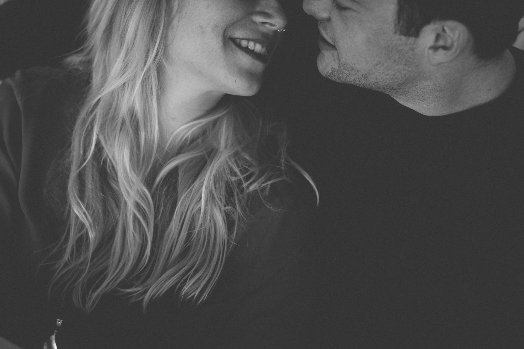 Nahaufnahme eines Paares, das seitlich face to face sitzt und sich anschaut und lächelt, die Frau hat lange blonde Haare. Schwarz-weiss-Aufnahme.