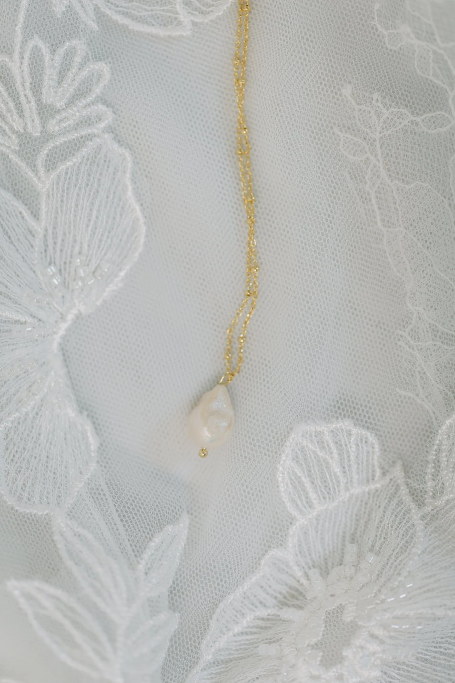 Goldkette mit einem tropfenförmigen Perlenanhänger auf weissem, transparentem Stoff mit floralem Stickmuster.