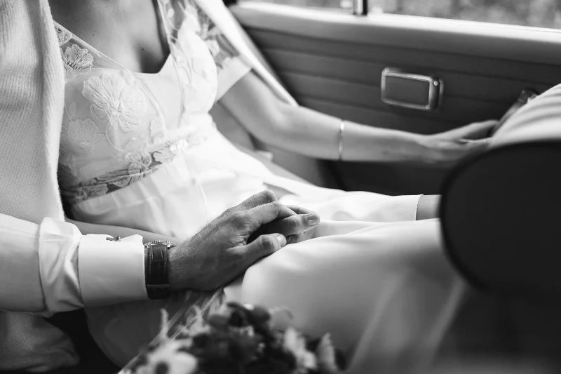 Hochzeitsfoto einer Braut und eines Bräutigams, er hält ihre Hand, sie sitzen im Auto.