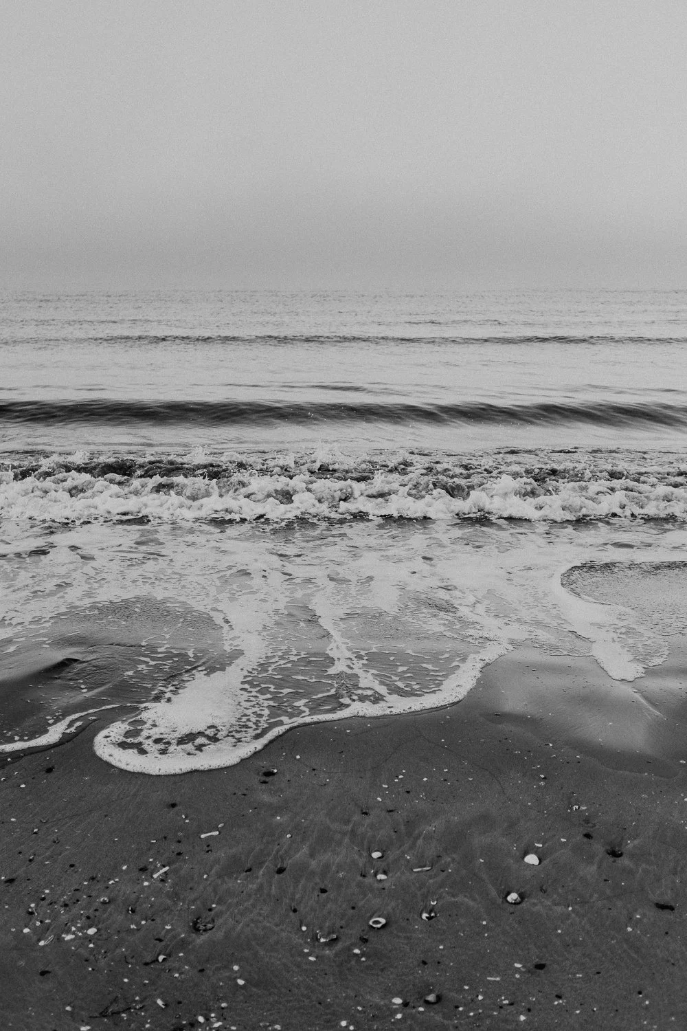 Schwarz-weiss-Foto eines ruhigen Meeres mit Wellen, die an den Strand spülen, und einem nebeligen Himmel im Hintergrund.
