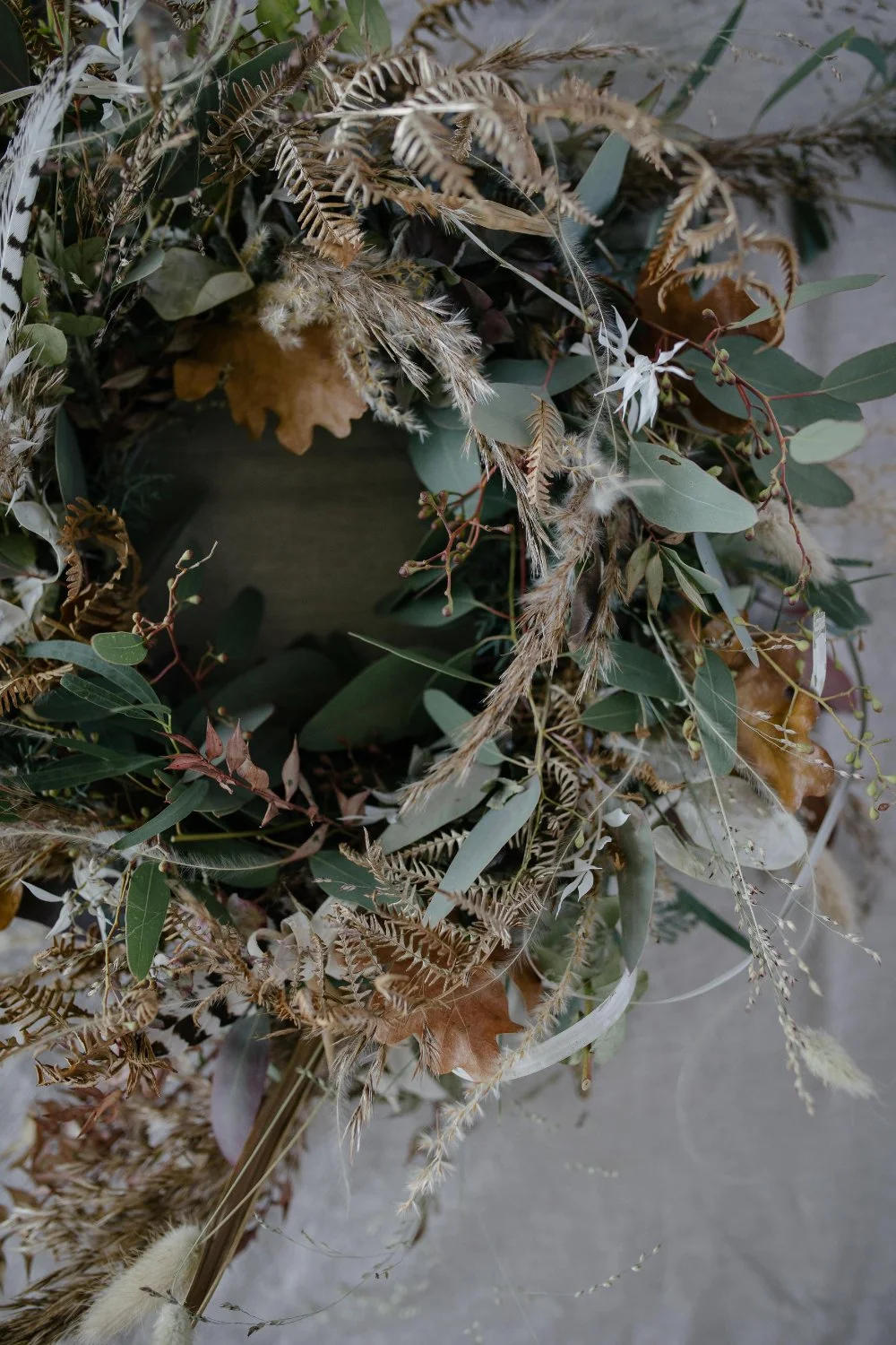 Ein rustikaler Blumenkranz aus getrockneten Blättern, Gräsern und Zweigen, teilweise mit weissen Blüten, auf einer grauen Oberfläche.