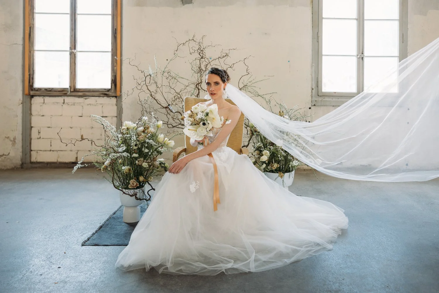 Eine Braut in einem weissen Brautkleid sitzt in einem verzierten Raum mit grossen Fenstern. Sie hält einen Blumenstrauss und trägt einen langen, fließenden Schleier. Der Hintergrund ist mit Blumen und Zweigen dekoriert.