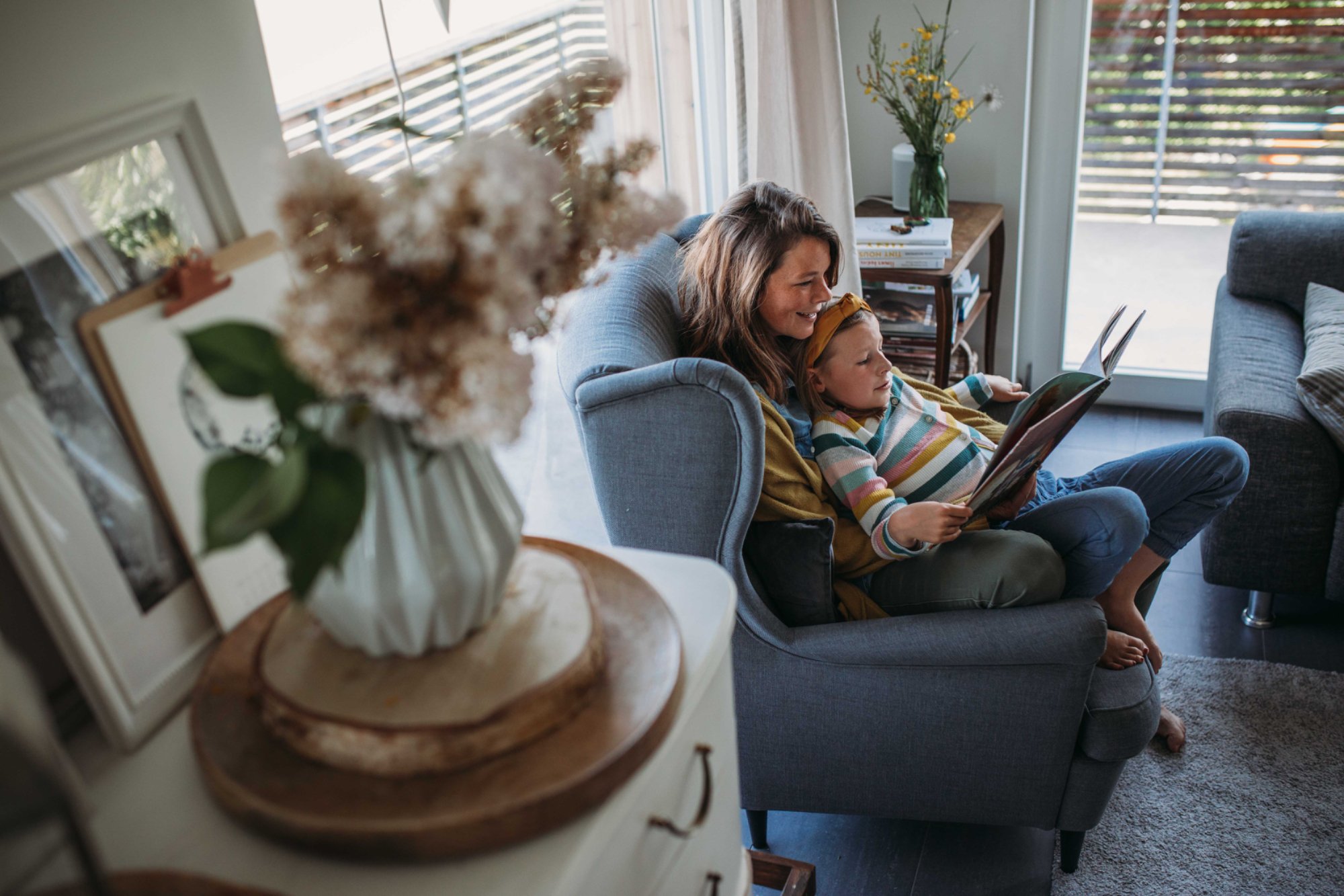 Eine Frau und ein Kind sitzen entspannt auf einem Ohrsessel und lesen gemeinsam ein Buch in einem Wohnzimmer mit grossen Fenstern und viel natürlichem Licht.