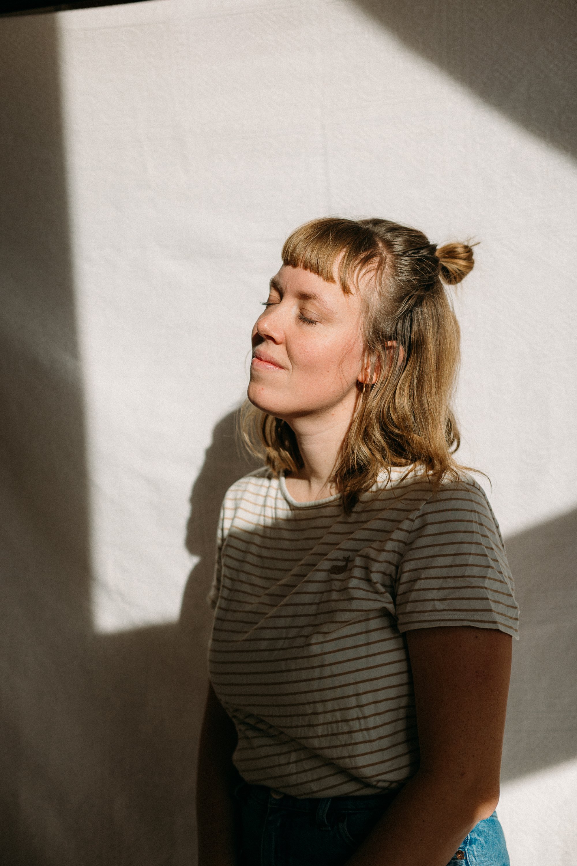 Junge Frau mit geschlossenen Augen, die Sonne scheint auf ihr Gesicht, während sie gegen eine weiße Wand lehnt.